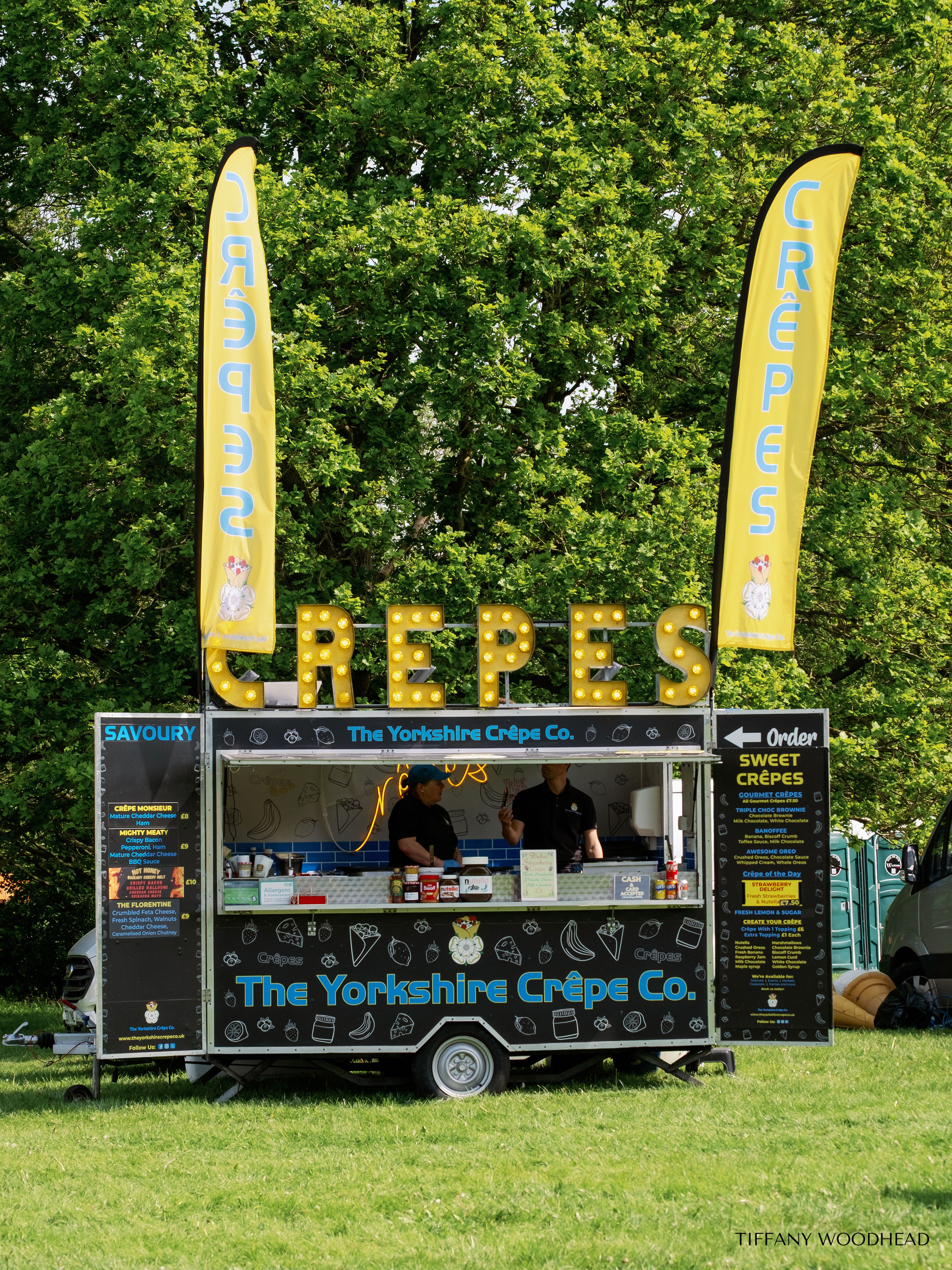 A mobile food stand for The Yorkshire Crêpe Co. decorated with black and blue, featuring yellow banners reading 'CRÊPES', with a menu on the side listing various sweet and savory crêpes, operated by two people wearing black shirts under a canopy with