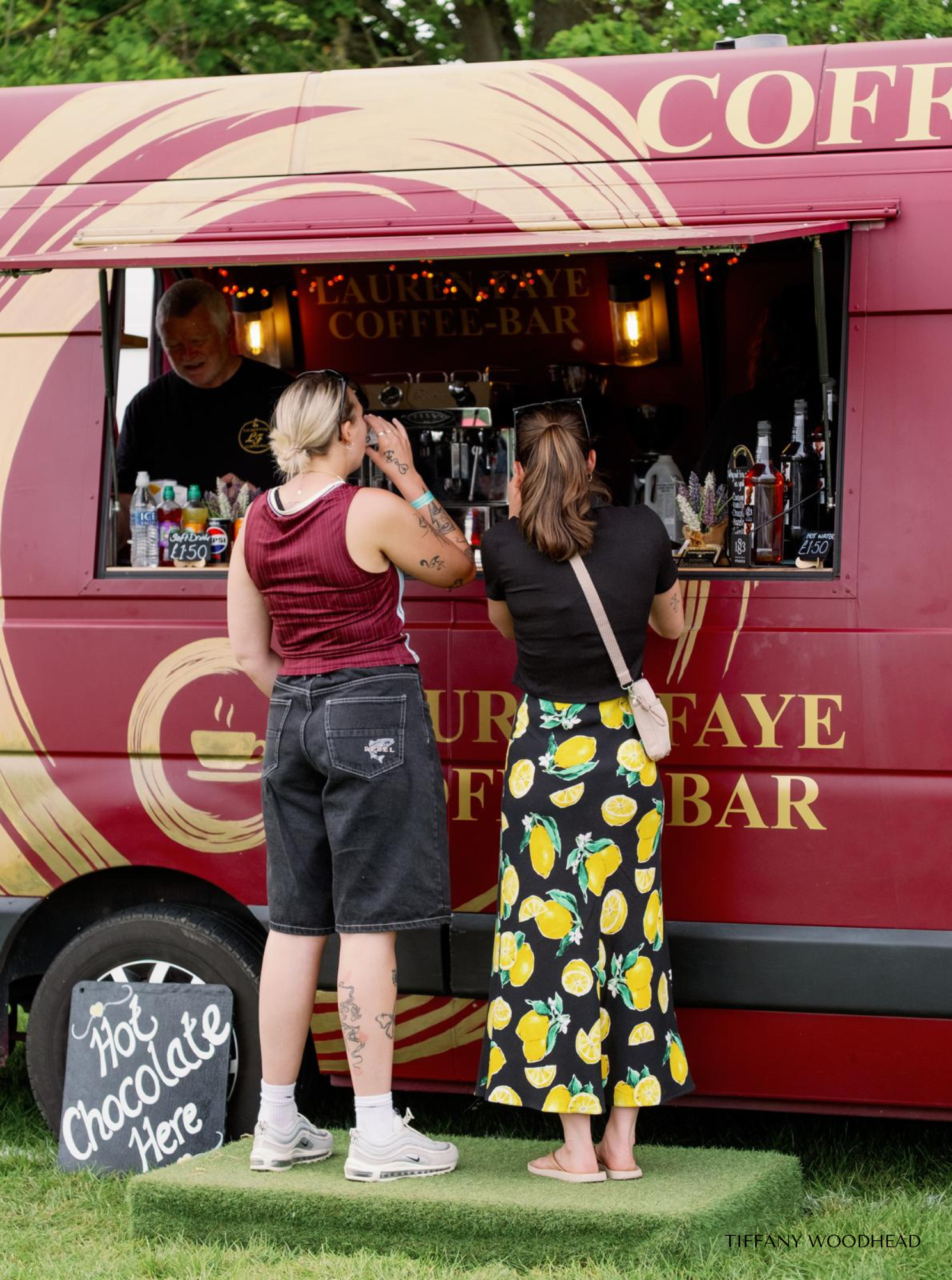 Two women ordering hot chocolate at a pink coffee stand decorated with signs and flowers, with a chalkboard sign in front that says 'Hot Chocolate Here.'