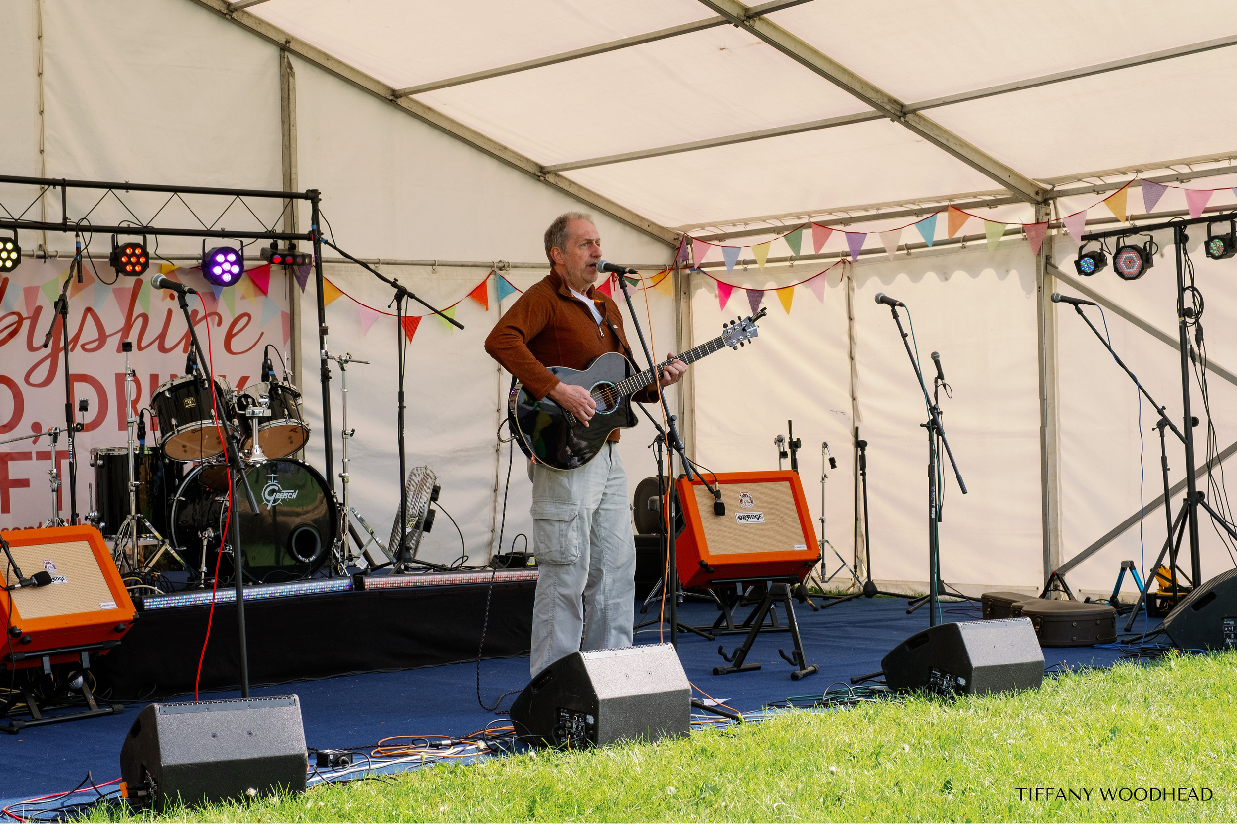 A man playing an acoustic guitar and singing into a microphone on an outdoor stage decorated with colorful bunting flags, with musical instruments and sound equipment around him.