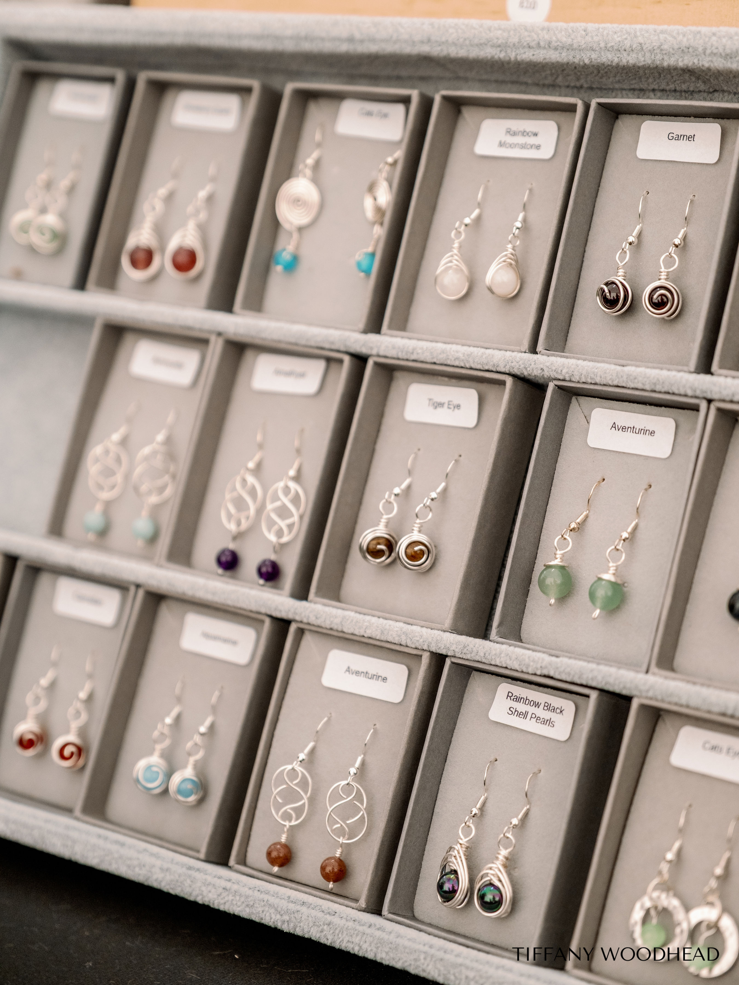 A display case with multiple rows of jewelry, including various earrings labeled with names like 'Rainbow Moonstone', 'Garnet', 'Tiger Eye', and 'Aventurine'.