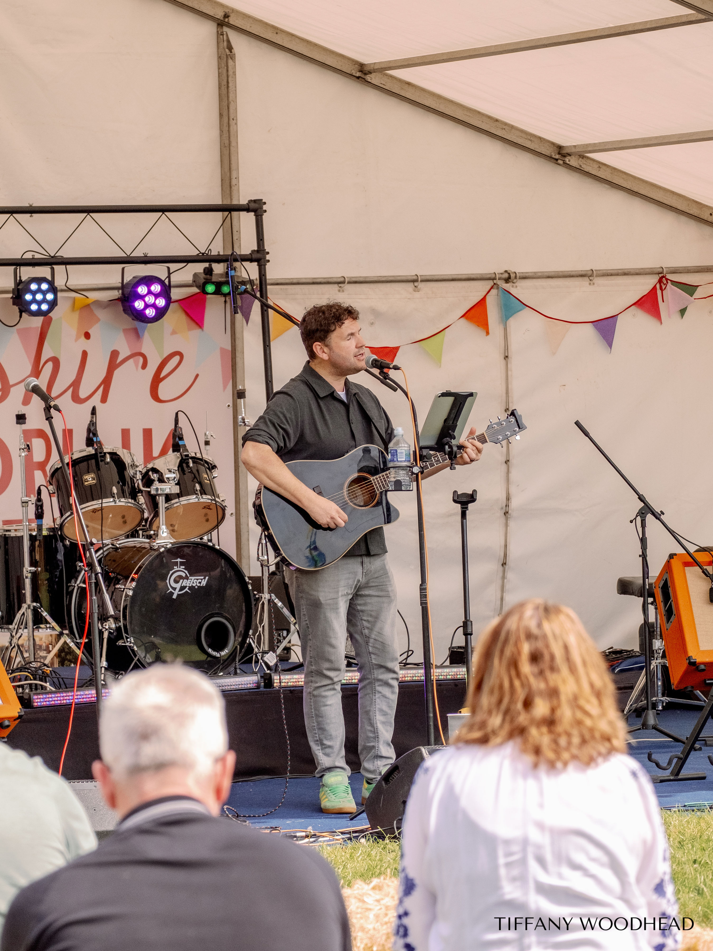A man with curly hair playing an acoustic guitar and singing into a microphone on a stage decorated with bunting and colorful lights, with an audience seated on the grass.