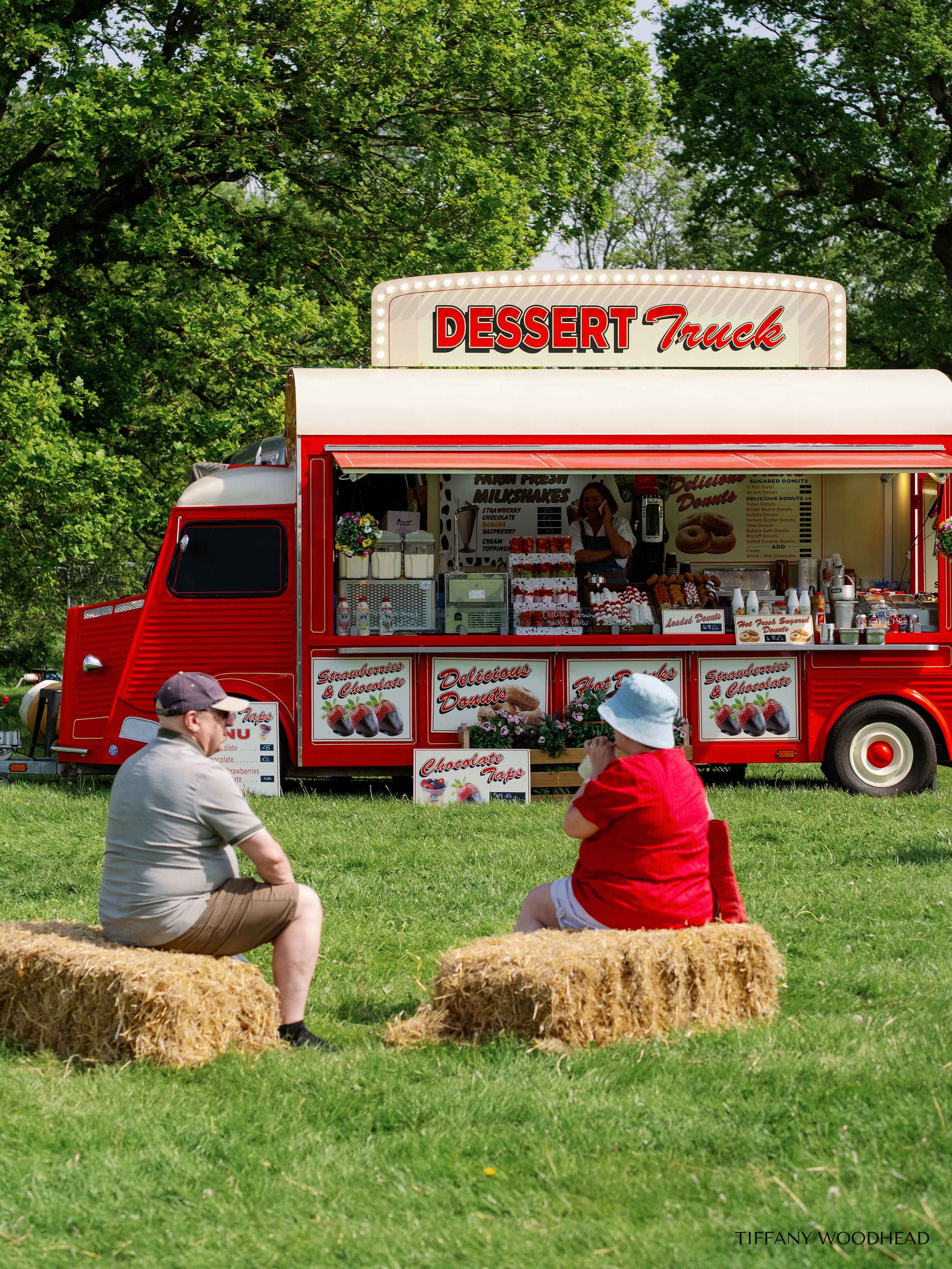 A red dessert truck selling donuts, strawberries, and chocolate treats at an outdoor event. Two people, a man and a woman, are sitting on hay bales in front of the truck, enjoying the day amid green trees.