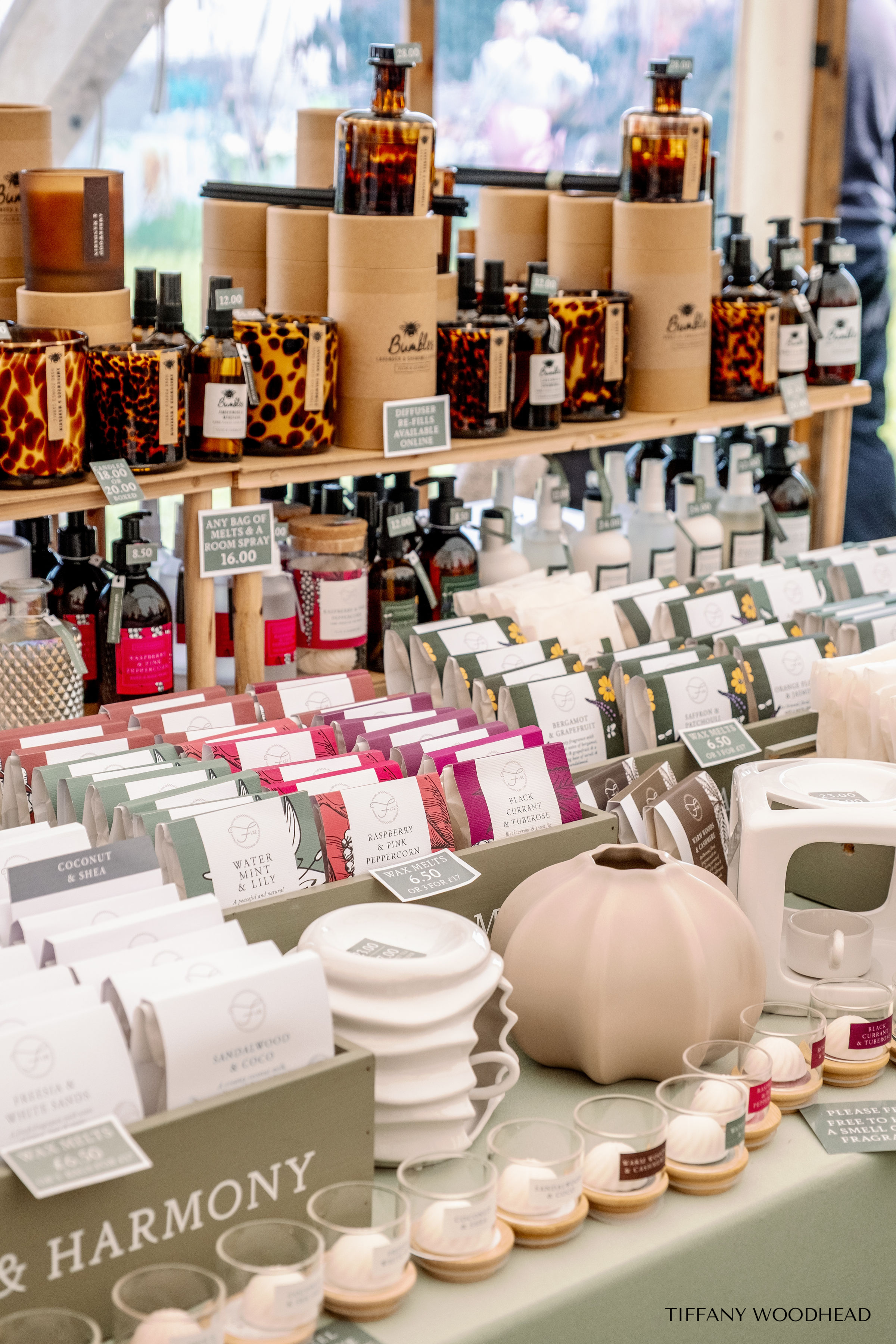 Display of candles, diffusers, and tea light holders on a wooden shelf at a market booth, with some about to be purchased.