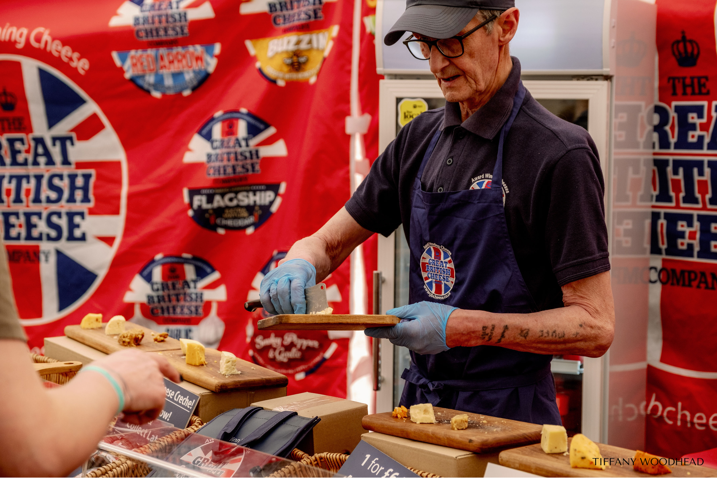 A man wearing a gray cap, glasses, and a navy apron, is serving cheese from a wooden board at a cheese booth with a red backdrop branding 'The Great British Cheese'.