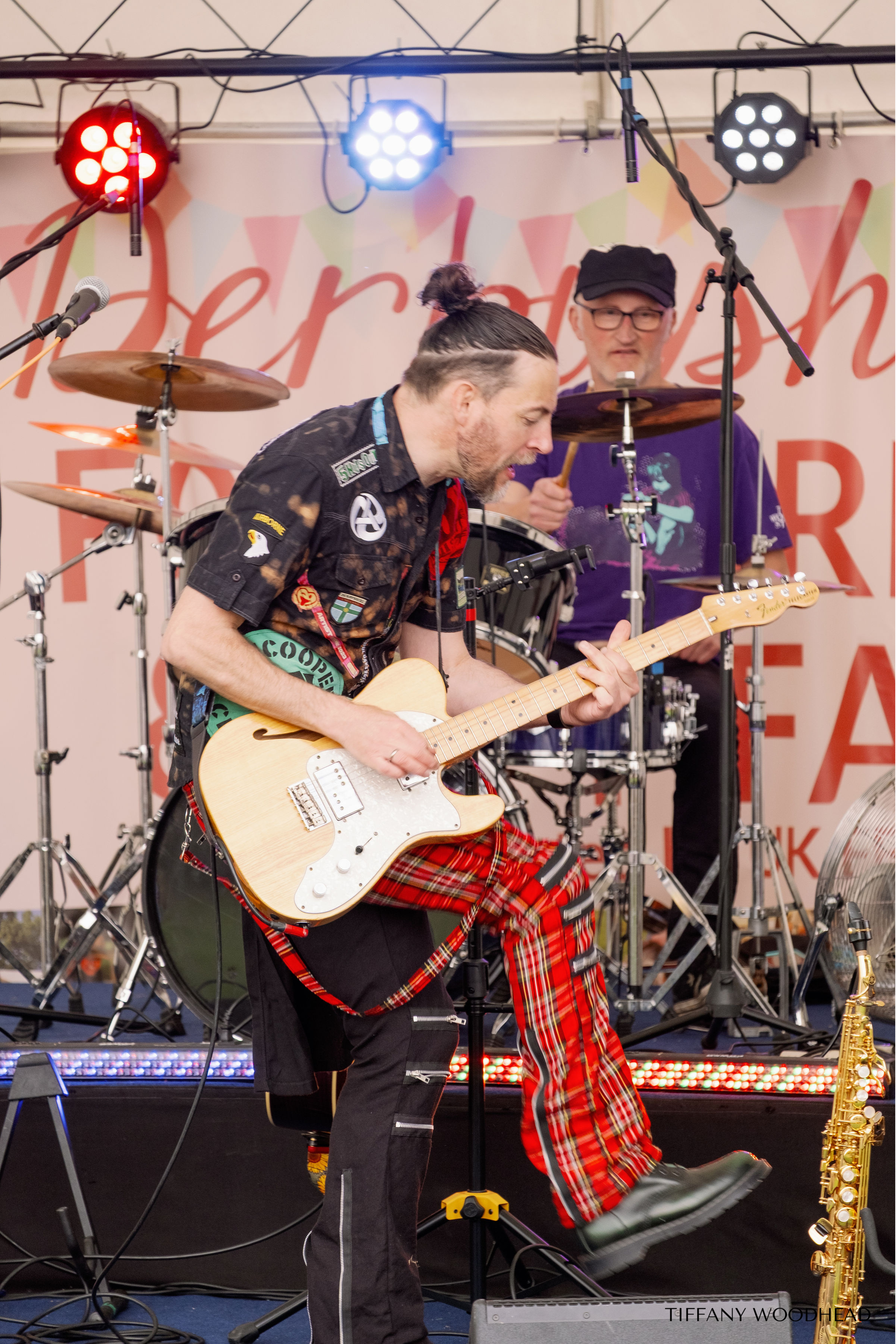 Two musicians playing instruments on stage at a live music event. The man in the foreground is playing an electric guitar and wearing a patch-covered shirt and tartan pants. The man in the background is behind a drum set, wearing glasses, a black cap