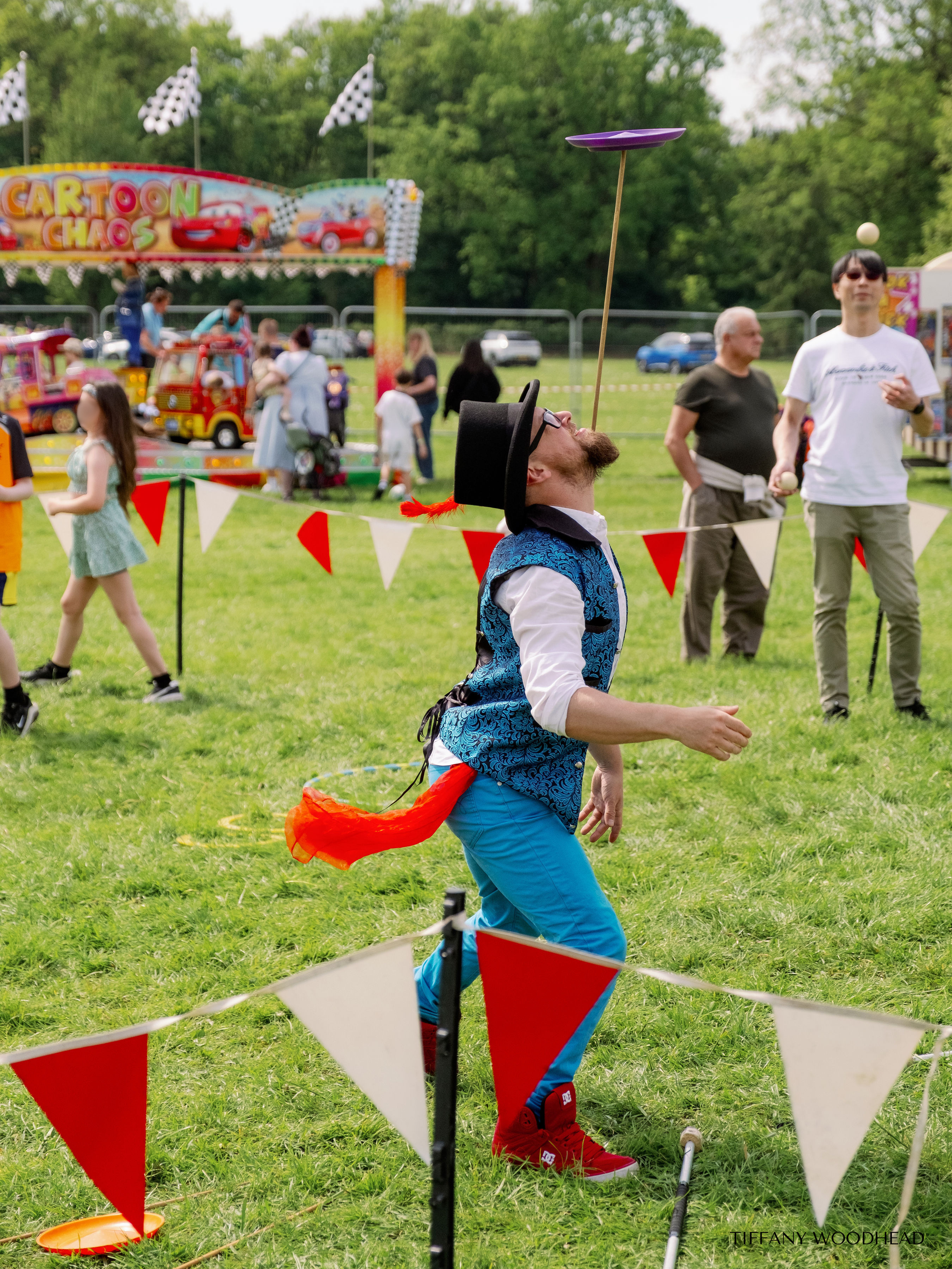 A man in colorful clothing and a hat is balancing a plate on a stick on his nose at an outdoor fair, with people and rides in the background.