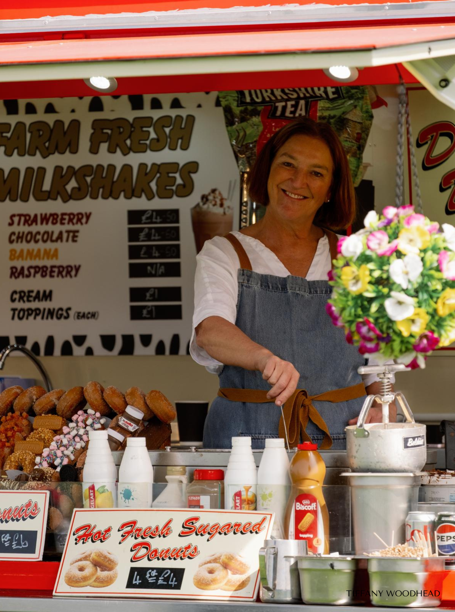 A woman at a food stand smiling, with a sign behind her advertising farm fresh milkshakes and donuts. The stand has various toppings, bottles of sauces, and a bouquet of pink and yellow flowers on the counter.