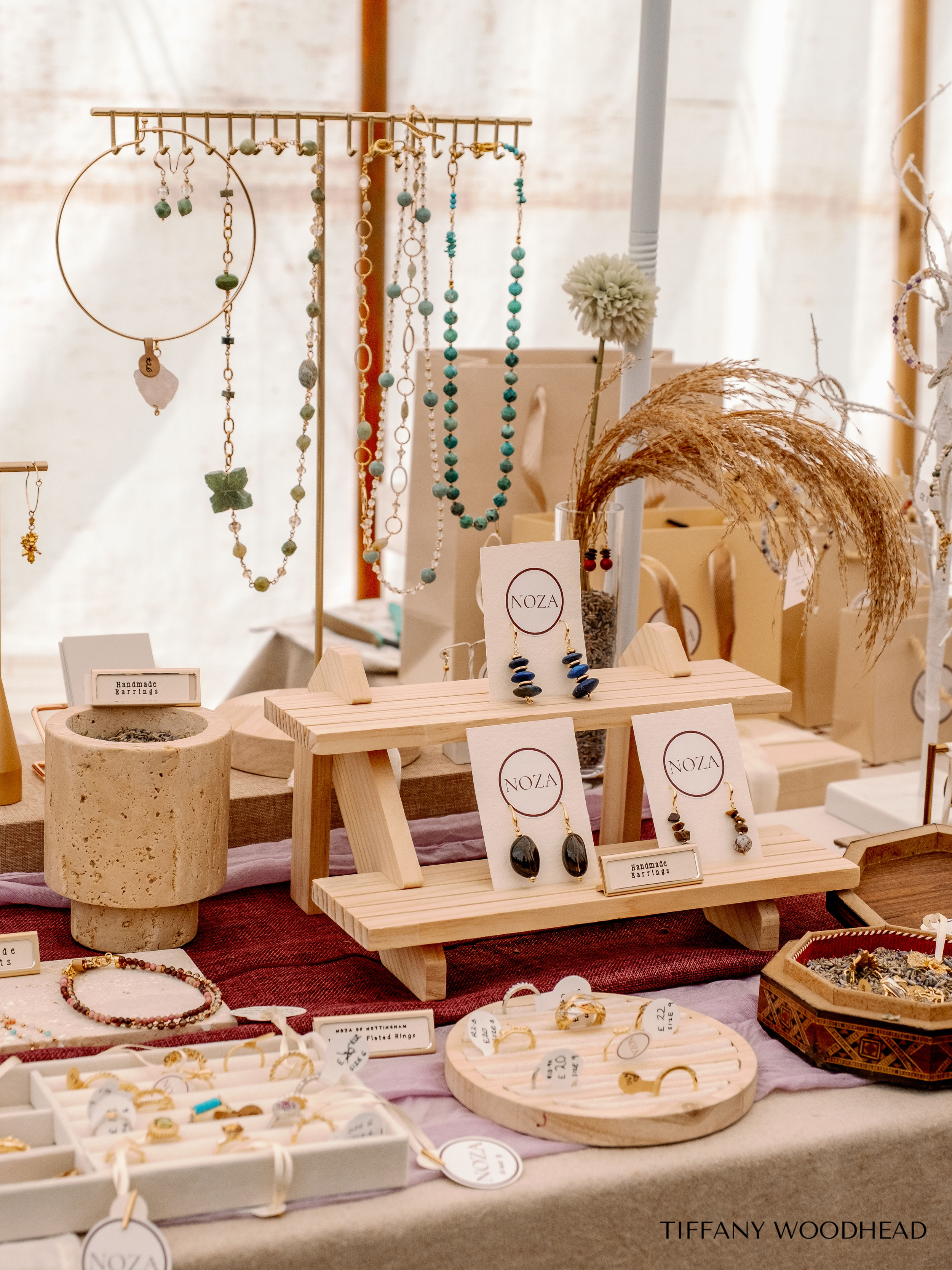 Display of handmade jewelry including earrings, necklaces, and rings arranged on wooden stands, trays, and tables at a craft fair, with some items labeled and a decorative dried plant piece on the right side.