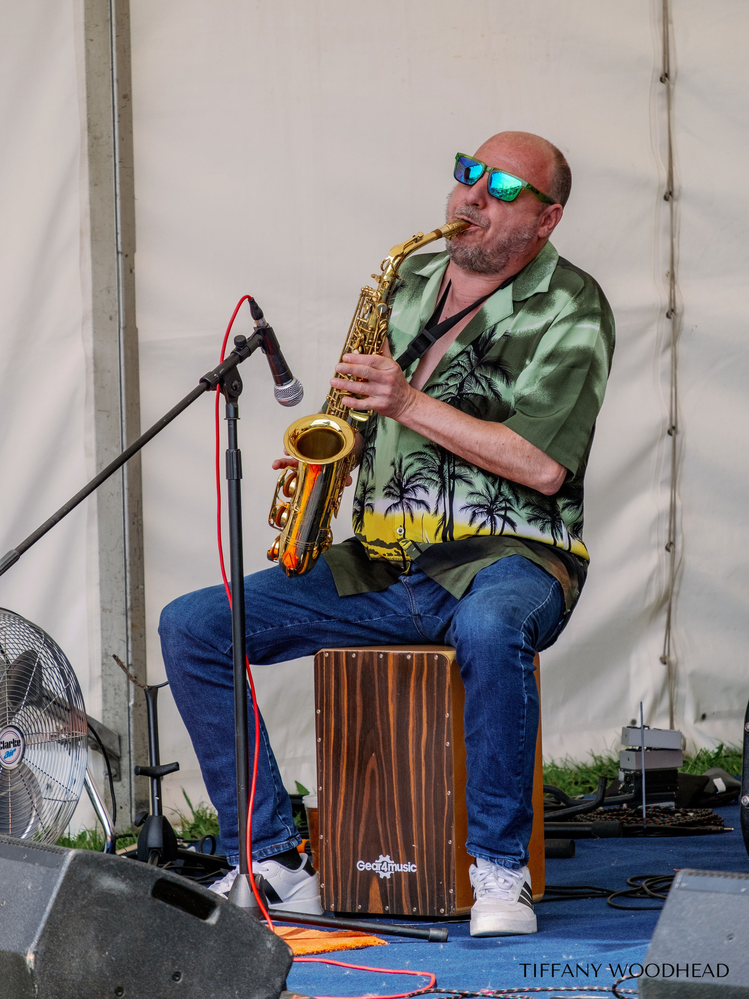 A man with sunglasses playing a saxophone while sitting on a cajón drum. He is wearing a tropical shirt and jeans, with a microphone in front of him. The background is a plain white tent wall, and there is audio equipment and a fan nearby.