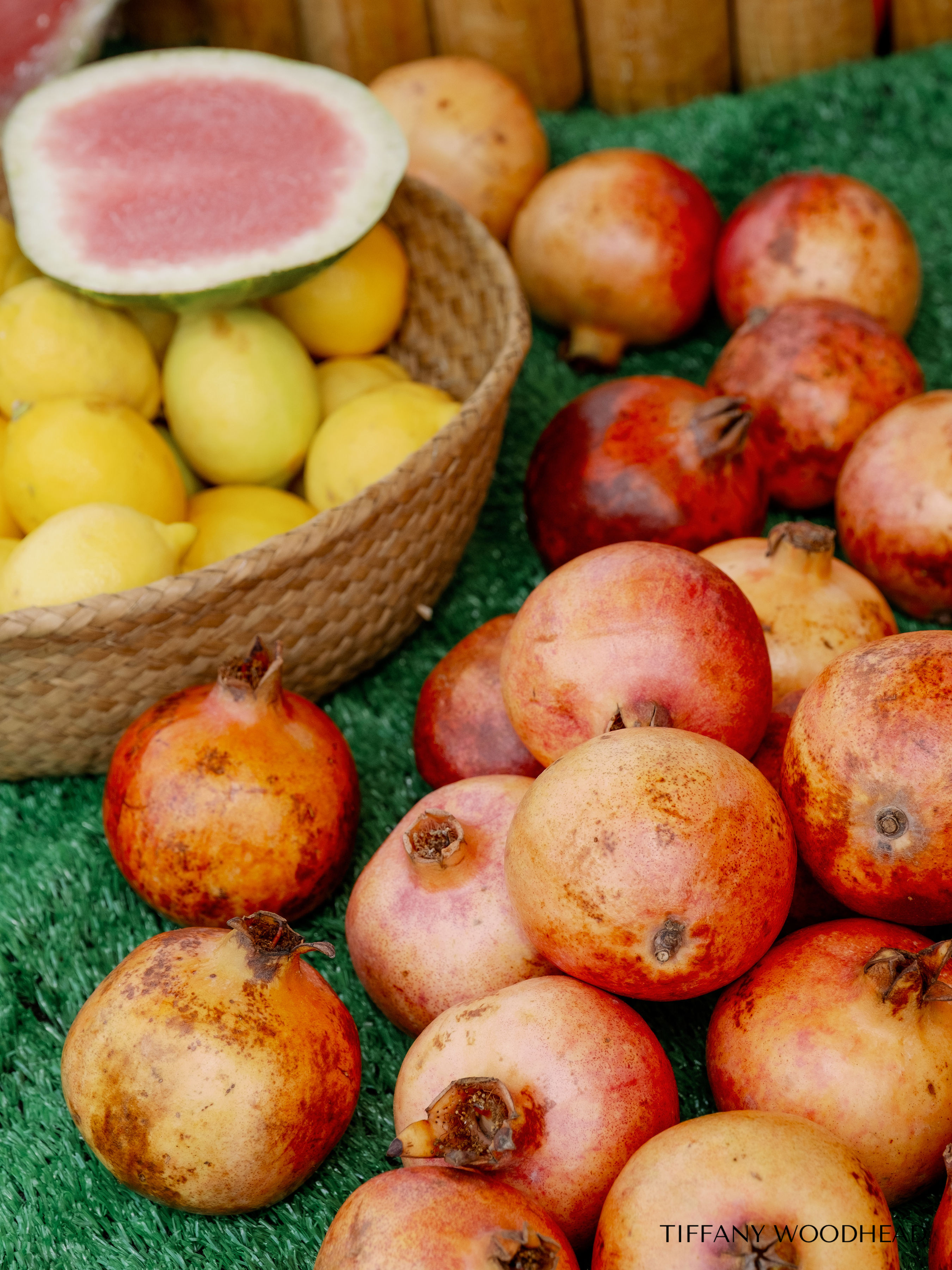 A display of pomegranates with pinkish-red and orange skin on a green surface, a basket of yellow quinces with a cut watermelon on top, and a wooden background.