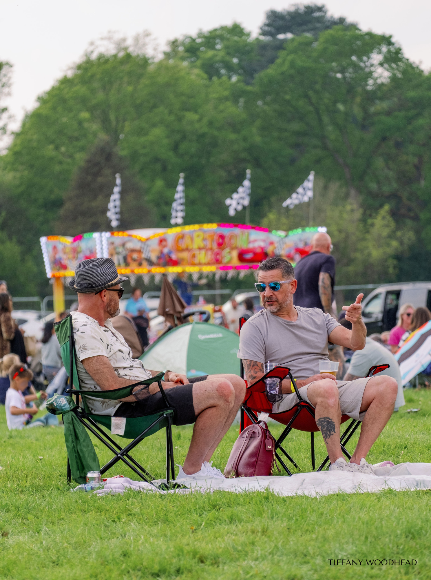 Two men sit in foldable chairs on a grassy field at an outdoor event. One man wears a striped hat and glasses, the other has sunglasses and tattoos on his arm, and is gesturing with his hand. There are tents, a carnival ride, and other people in the 