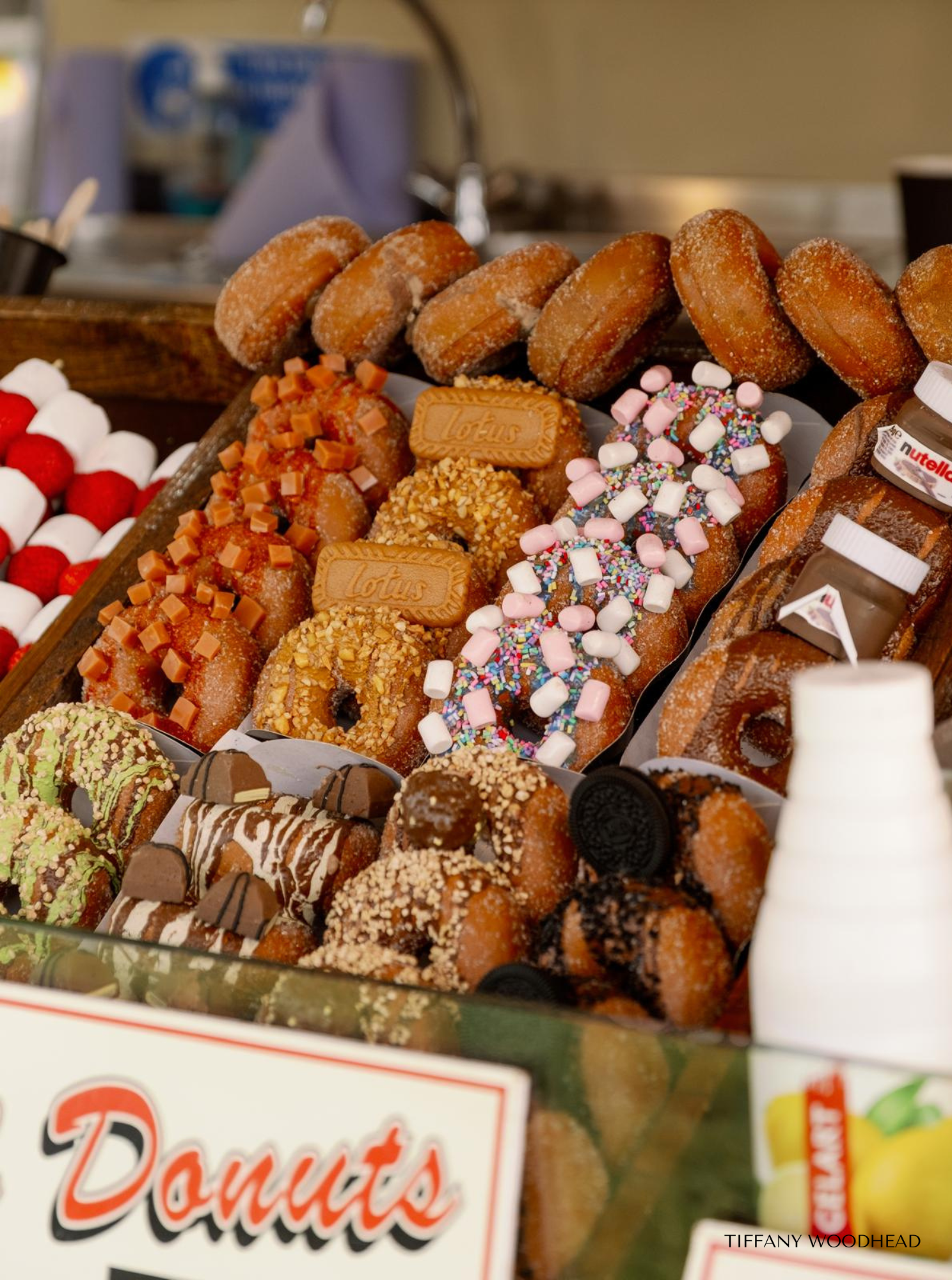 Assorted donuts decorated with various toppings including caramel, sprinkles, marshmallows, and chocolate, arranged in display trays.