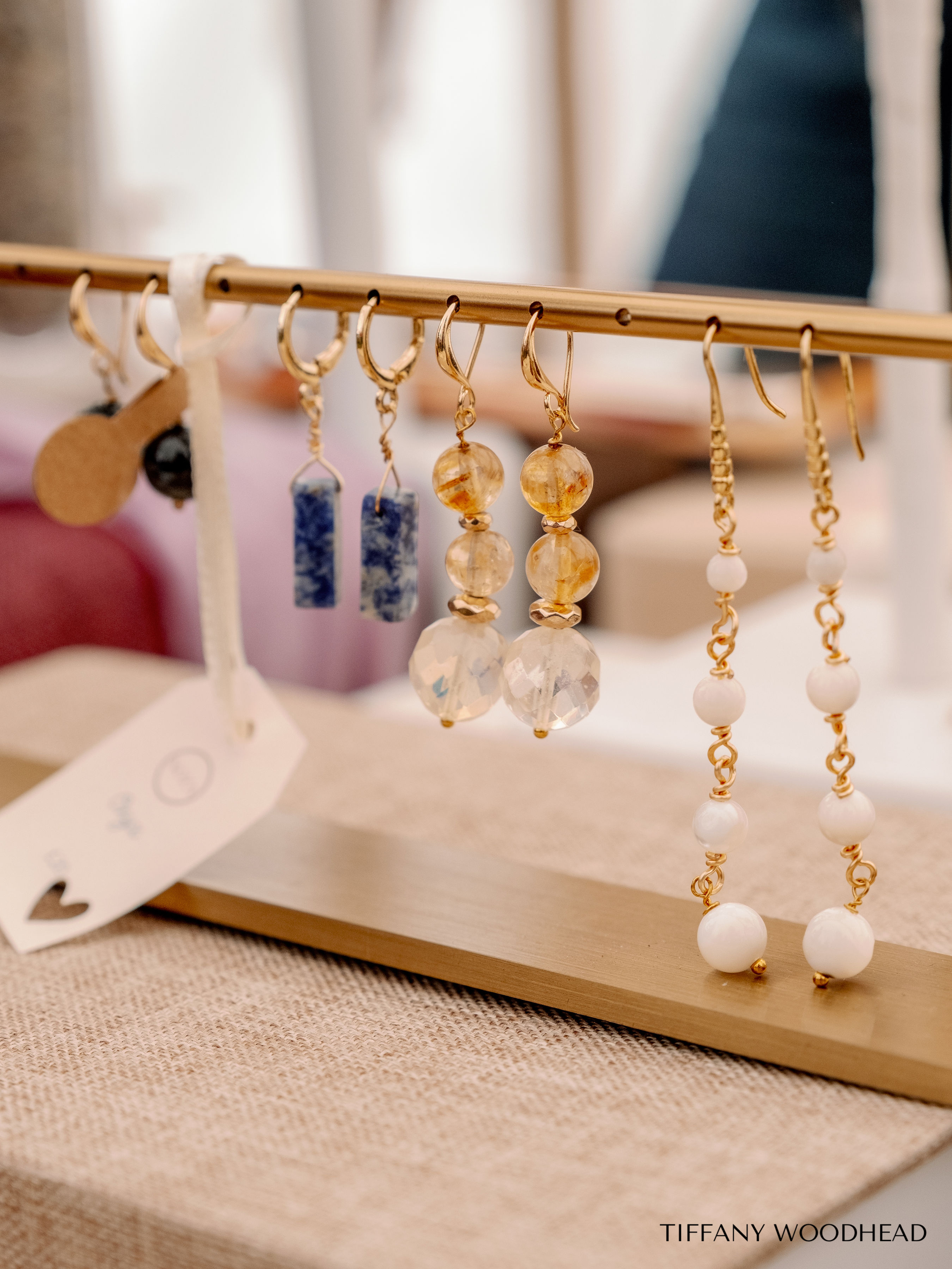 Close-up of a display of gold and beaded earrings hanging on a wooden stand on a beige fabric surface with Tiffany Woodhead's name at the bottom right.