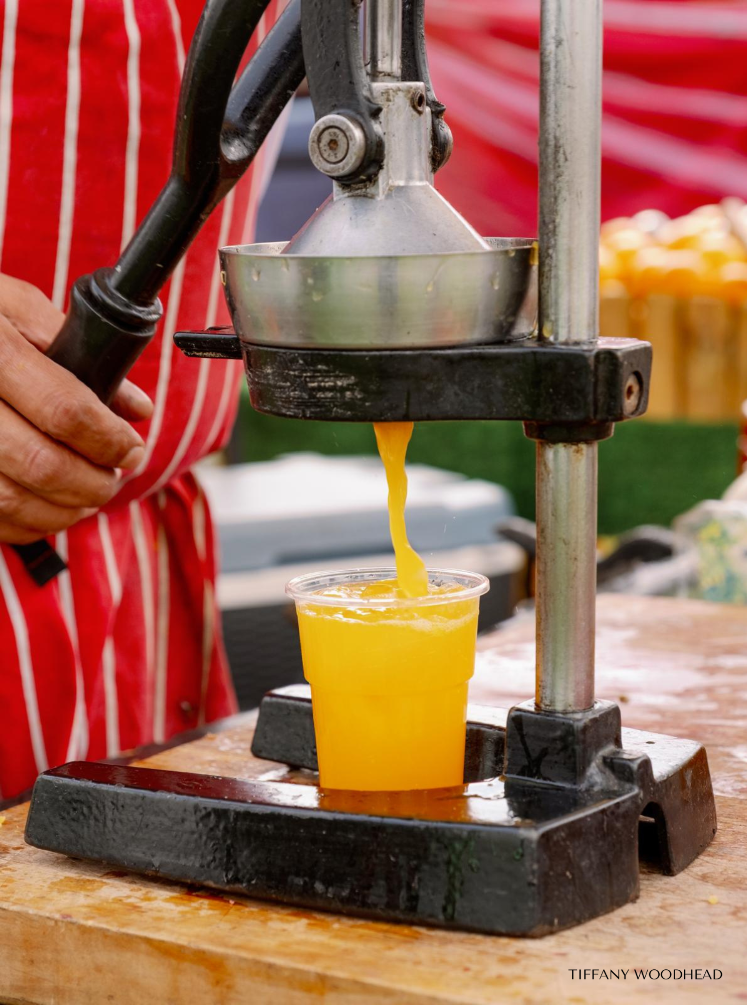 A person with a red and white striped shirt uses a manual juicer to extract juice from a yellow fruit, pouring the juice into a clear plastic cup. The juicer is on a black base on a wooden surface, with a red striped cloth in the background.