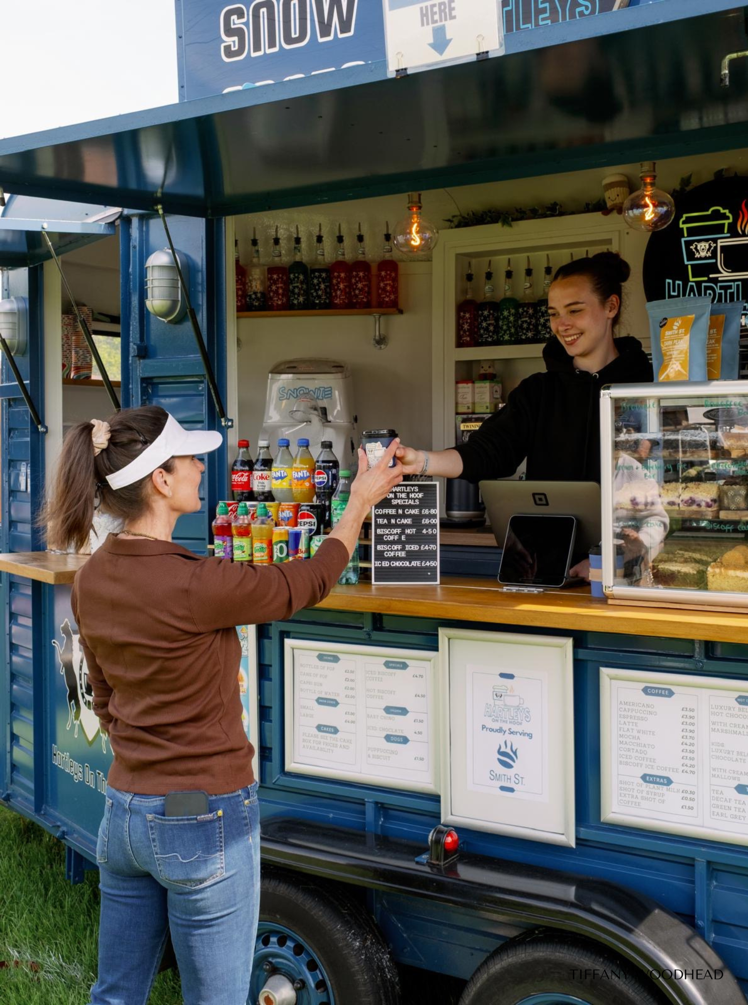 A woman with a white visor and brown sweater is buying a drink from a smiling girl working at a blue food truck. The truck has a menu and soda bottles on the counter, and the background shows shelves with cups and supplies.