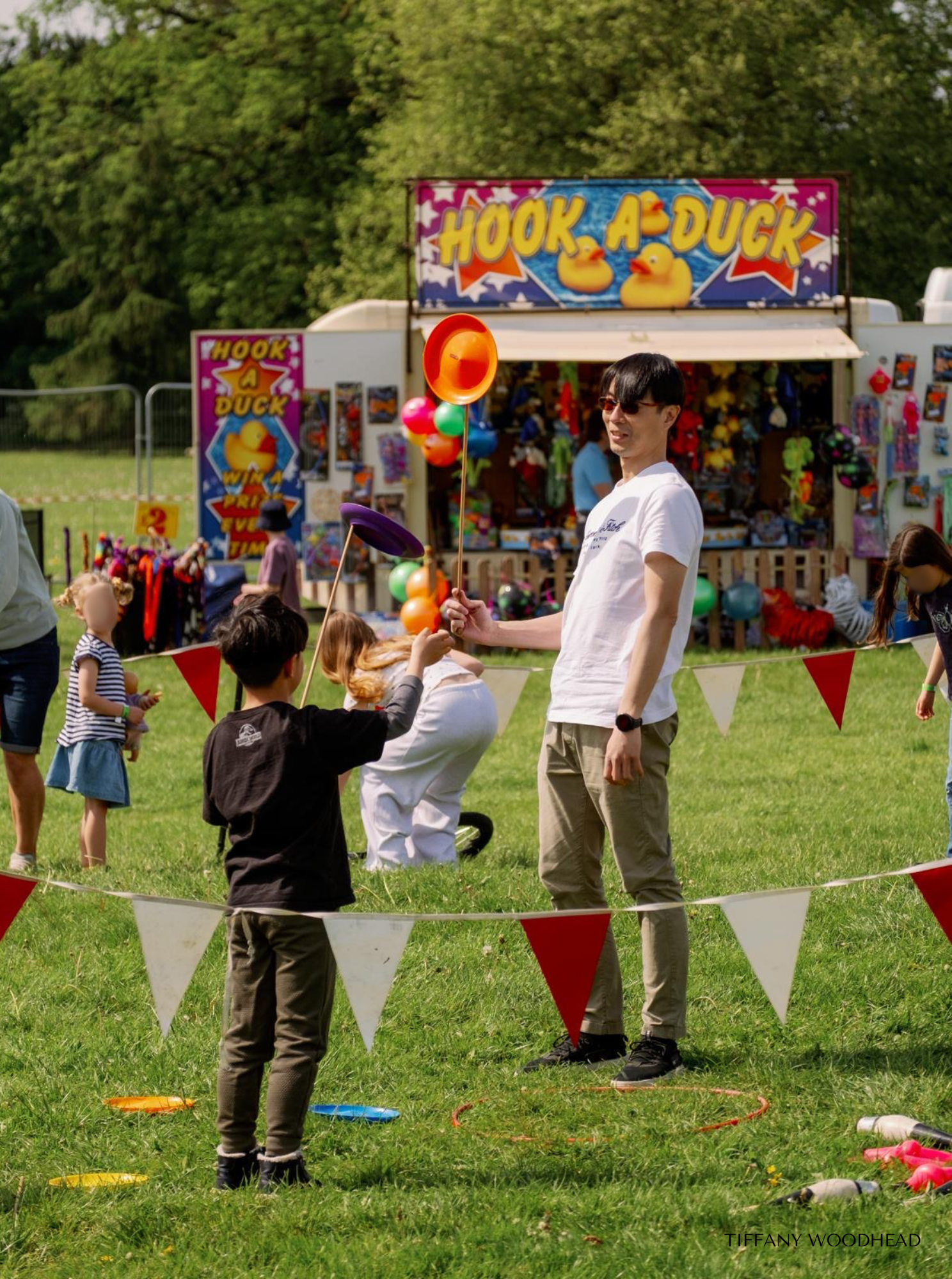 A man and a child playing a ring toss game called "Hook a Duck" at an outdoor fair. The game booth features colorful balloons and a sign with rubber ducks. People, trees, and other fair attractions are visible in the background.