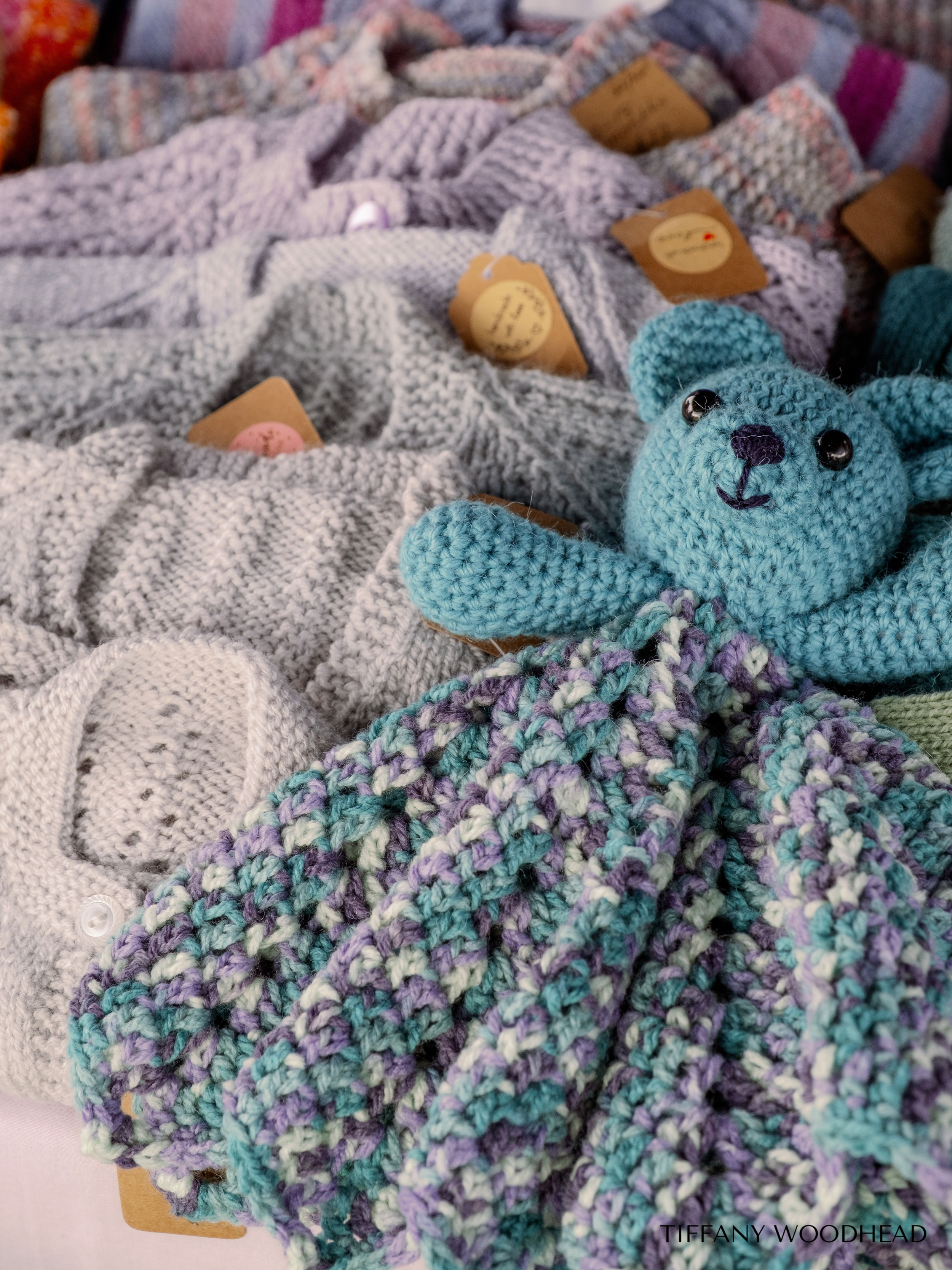 Display of various knitted scarves and a plush blue teddy bear with black eyes and a purple nose, arranged on a surface. Some scarves have tags attached.
