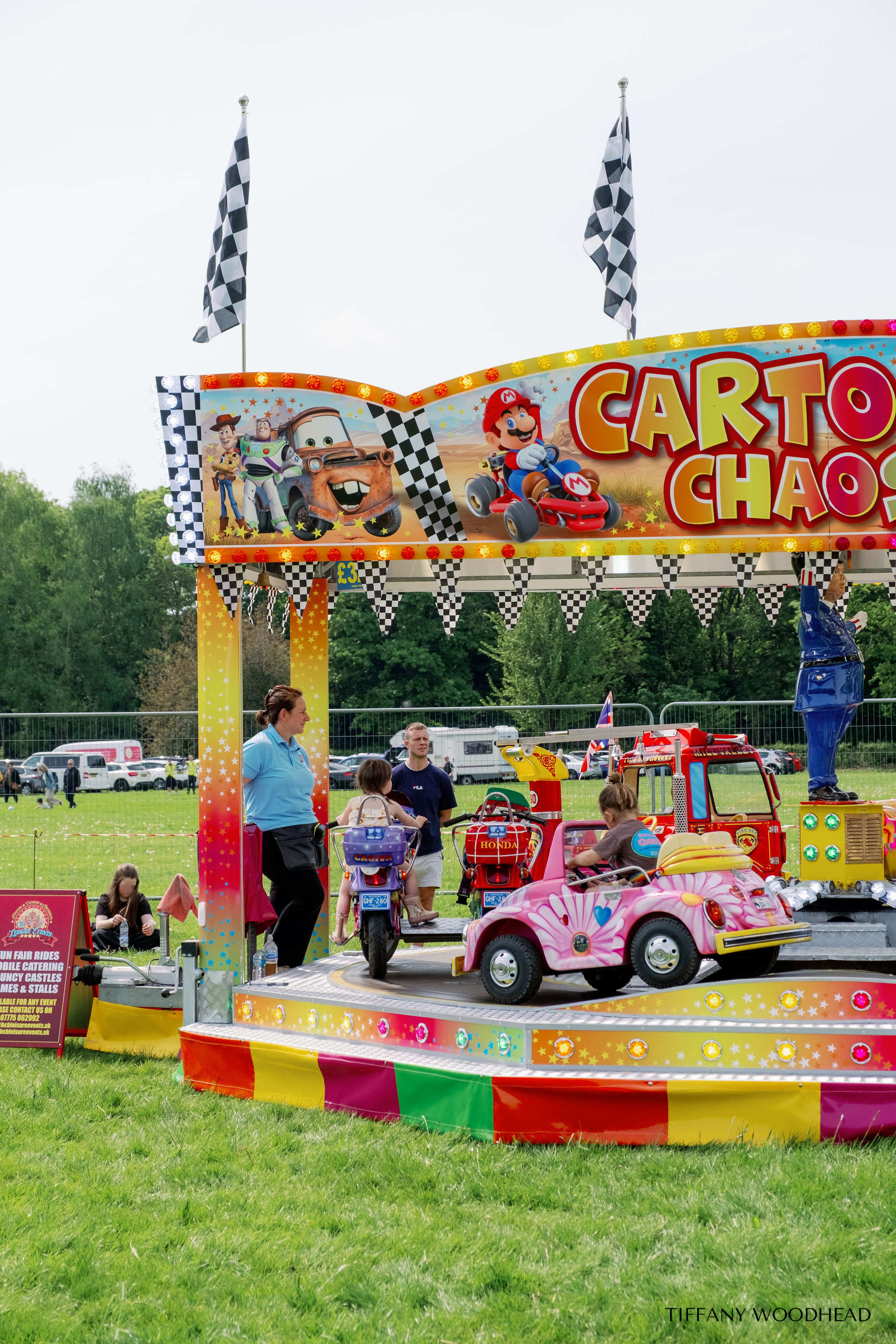 Colorful amusement ride with cartoon characters from Toy Story and Mario; children riding small cars, adults supervising; outdoor fair setting with green grass and trees.