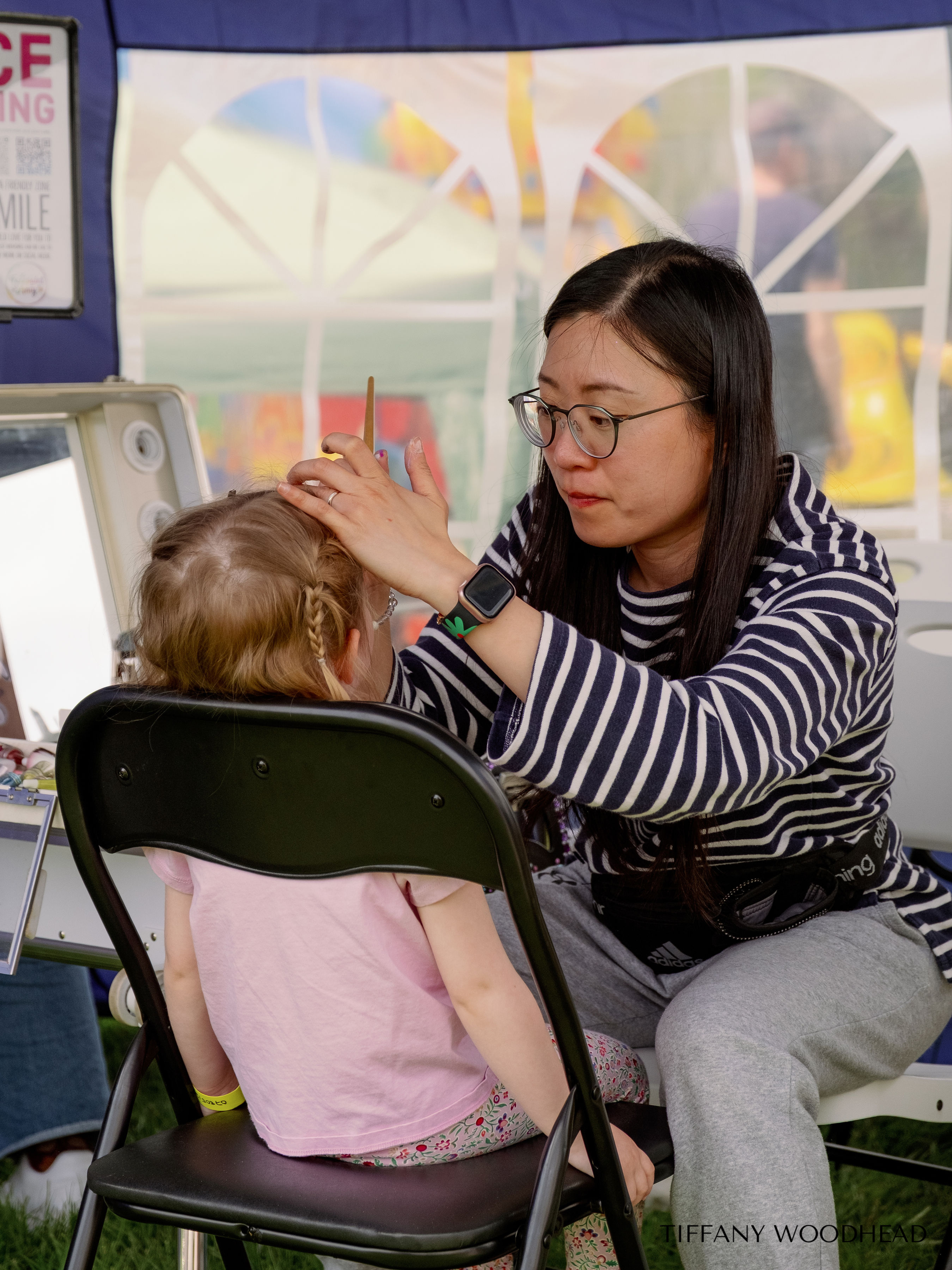 A woman with long dark hair and glasses, dressed in a striped shirt and gray sweatpants, is sitting on the ground in front of a young girl with braided blonde hair, wearing a pink shirt and floral leggings. The woman appears to be drawing or applying