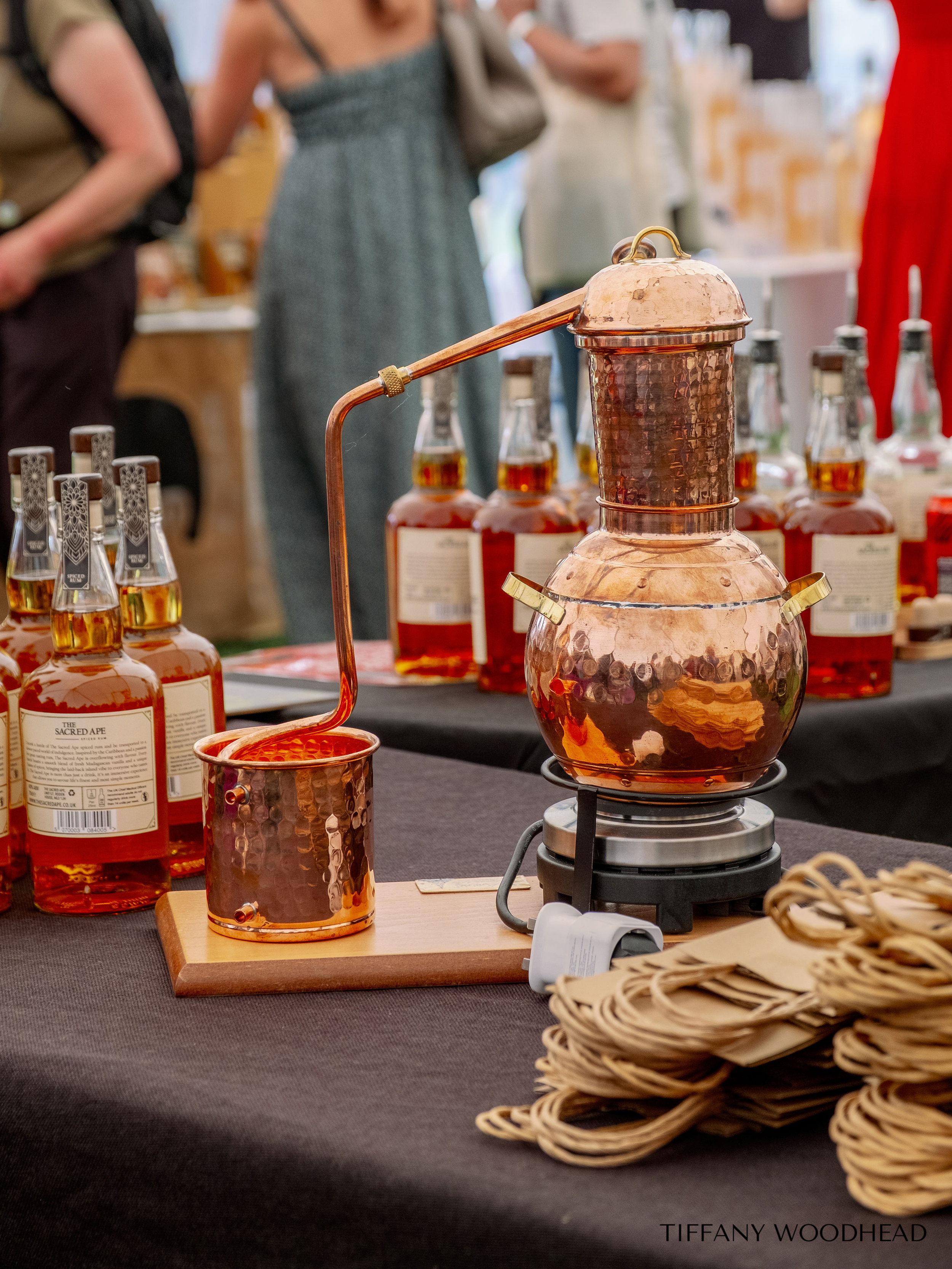 A copper moonshine still with a copper coil, a copper collecting cup, and bottles of liquor on a black tablecloth at an event. There are blurred people in the background.