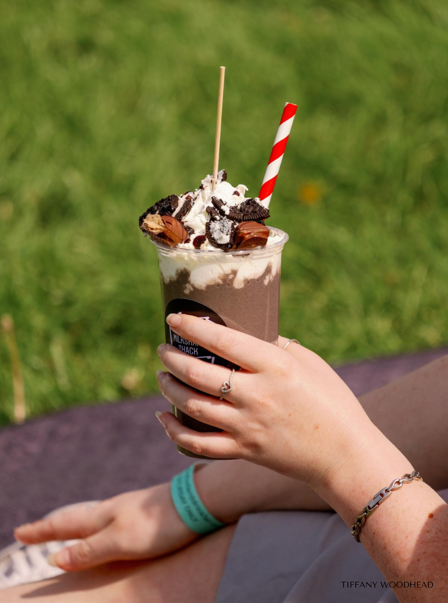 A person's hand holding a chocolate milkshake topped with whipped cream, Oreo cookies, and chocolate pieces, with red and white striped straw and wooden toothpick, outdoors with grassy background.