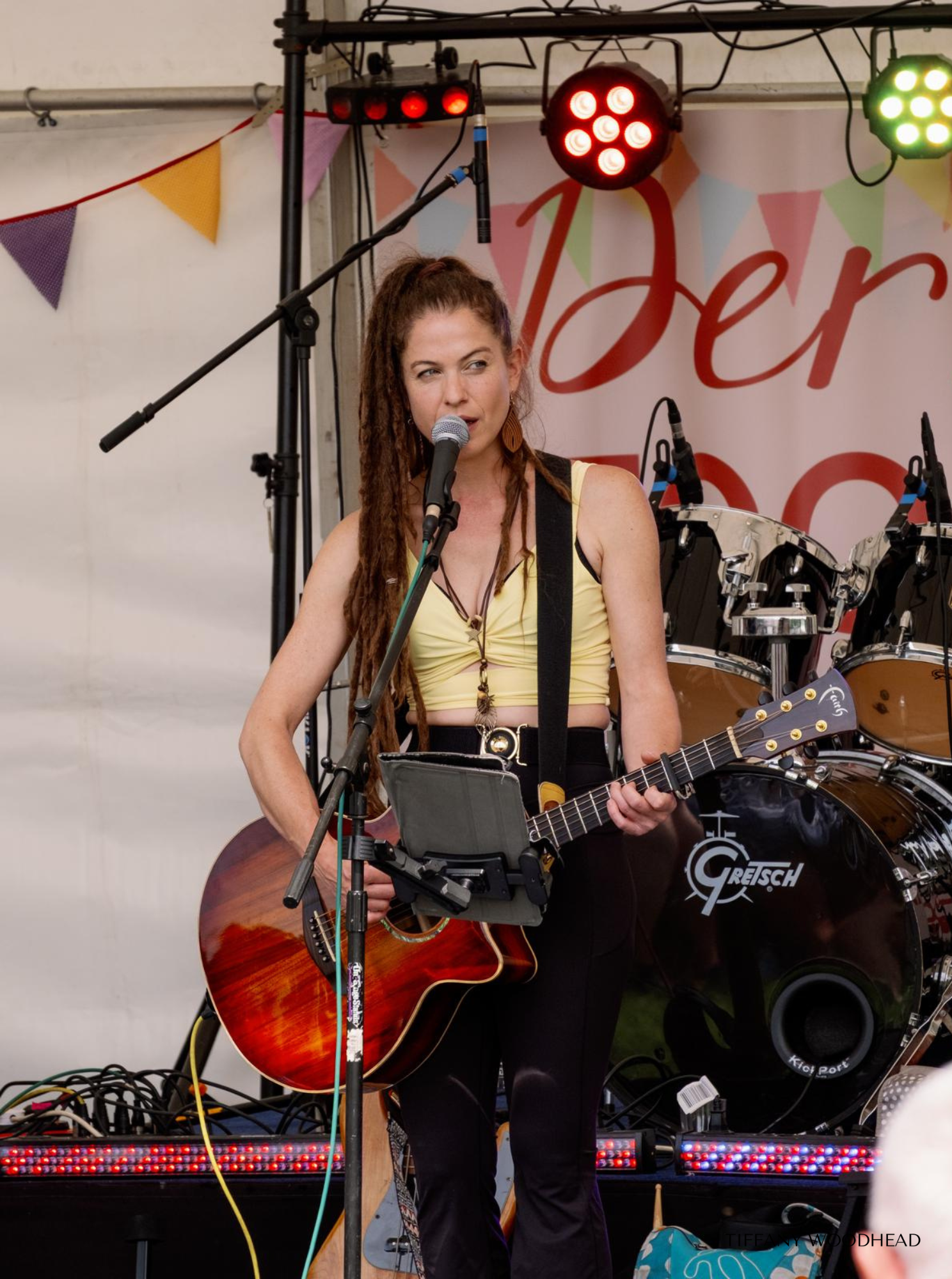 A woman with long dreadlocks performing on stage, holding a guitar and singing into a microphone. Colorful stage lights and a drum set are visible in the background.