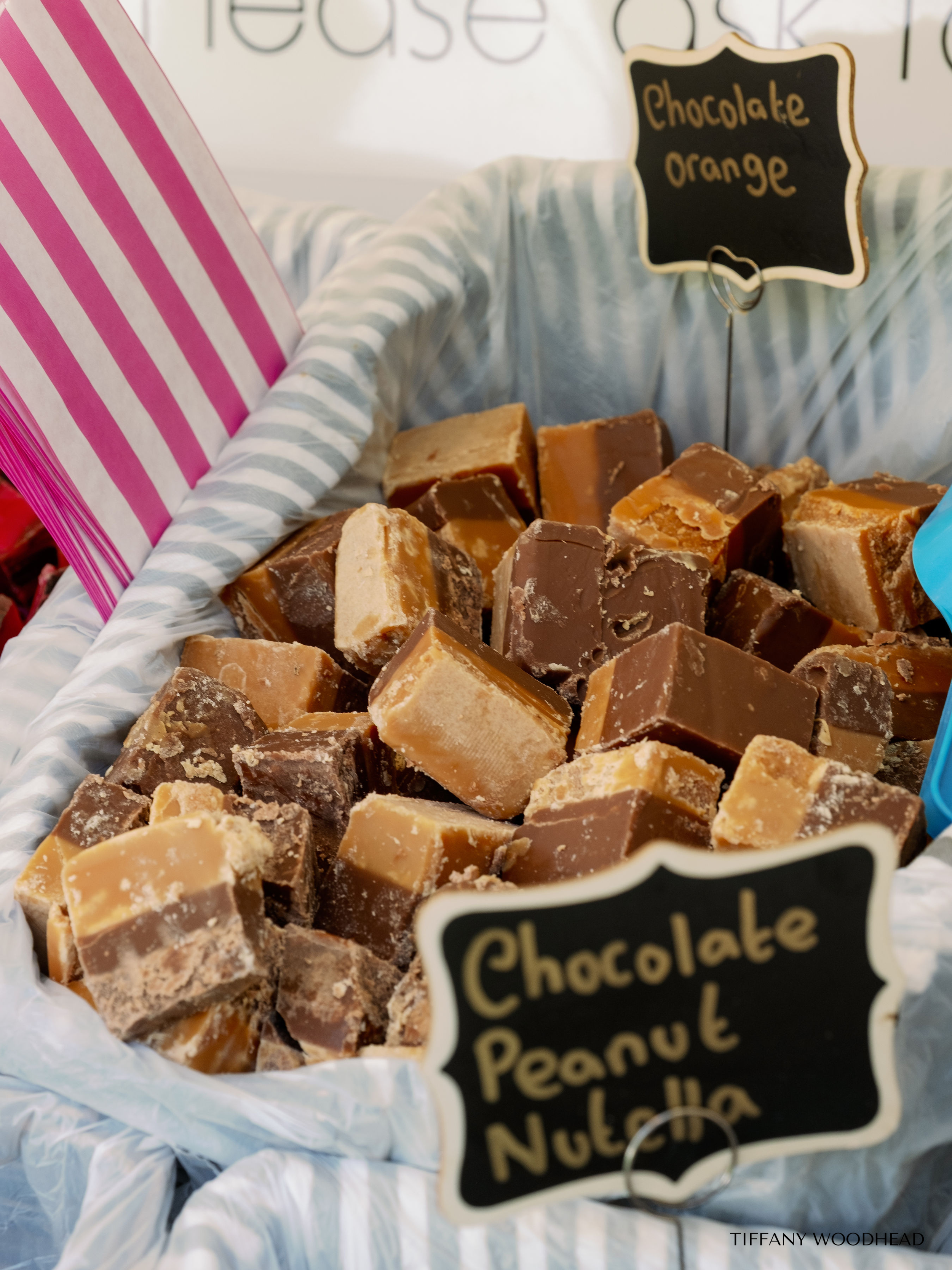 A basket of assorted chocolates and caramel fudge chunks, with a sign reading "chocolate orange". The basket is lined with striped tissue paper, and there is a small chalkboard label in the foreground with the sale handwritten below the chocolates.