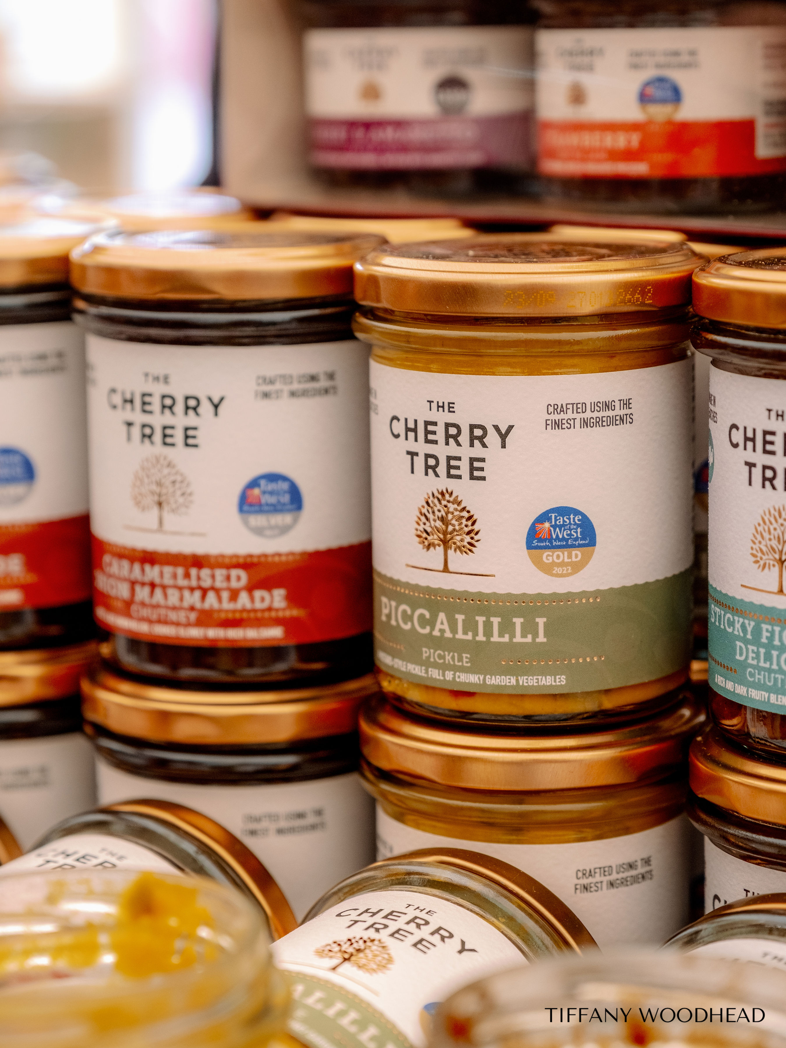 Close-up of jars of The Cherry Tree brand preserves on a store shelf. The labels display the brand name, product name, and a picture of a tree, with different flavors visible. The jars have gold lids and are stacked in multiple rows.