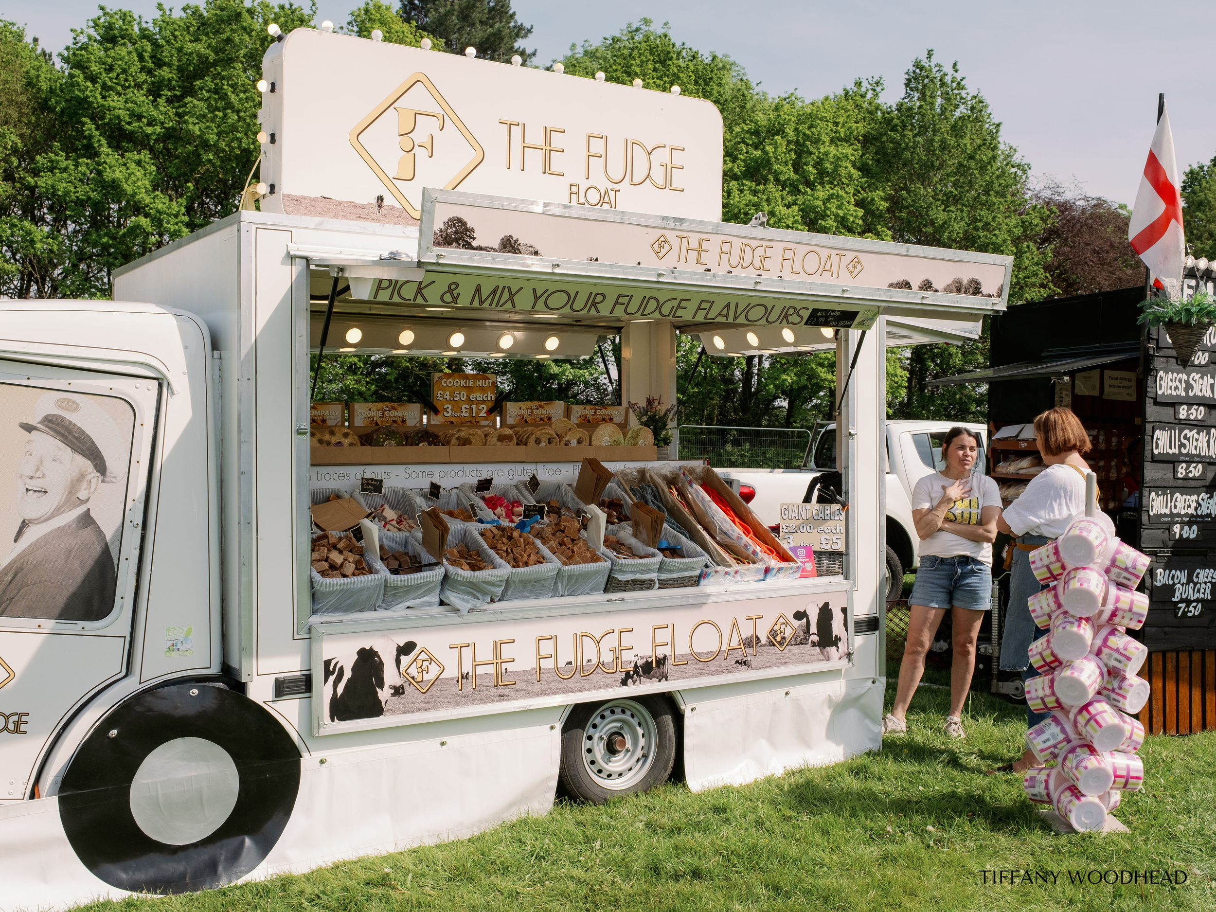 A mobile food stand titled 'The Fudge Float' offers various fudge flavors and treats at an outdoor event. The stand has a sign with a logo and the name, with tables of fudge blocks for sale. Two women are talking near the stand, with a stack of pink 
