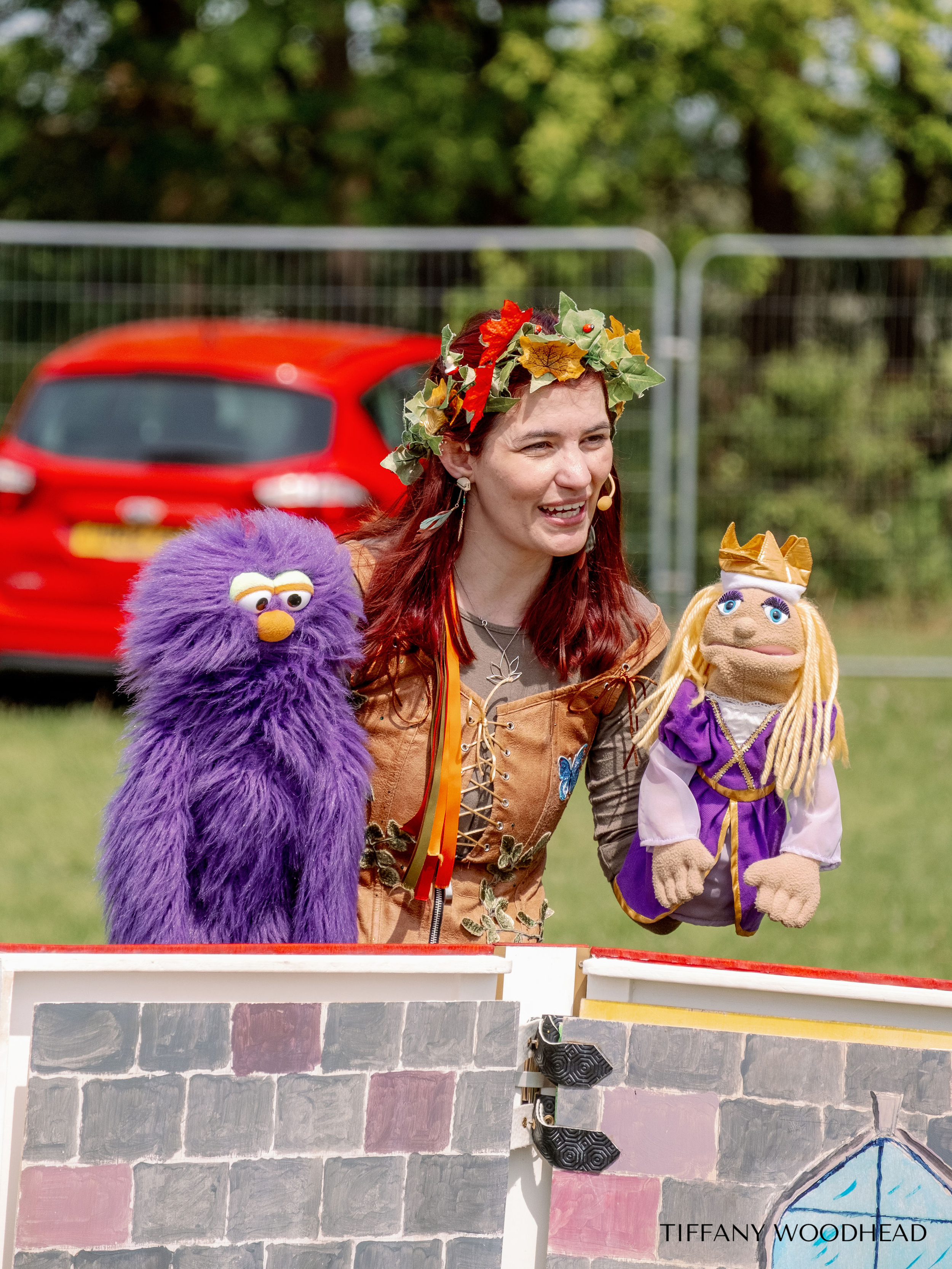 A woman with red hair wearing a leafy crown, smiling while holding puppets, standing outdoors behind a painted brick wall in front of a metal fence and green trees.