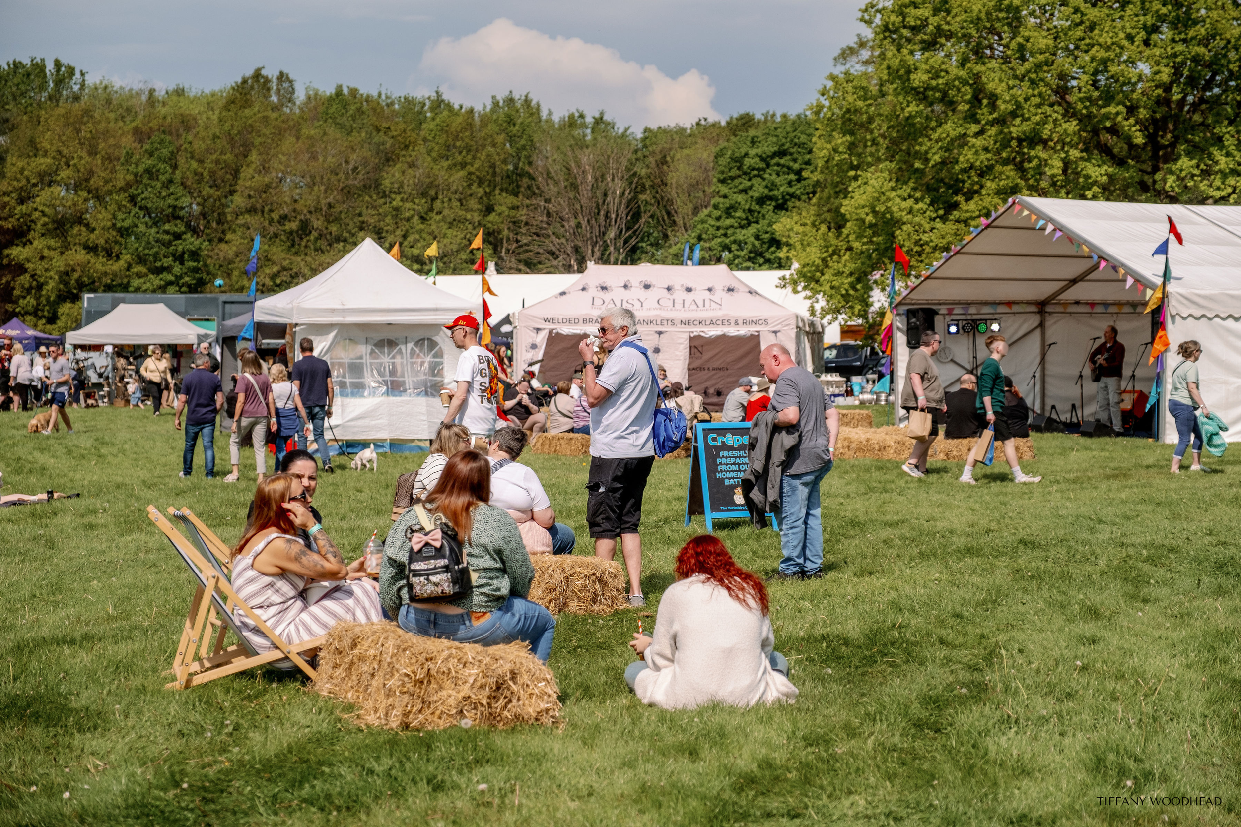 People attending an outdoor festival with tents, a stage with musicians, hay bales, and trees in the background during daytime.