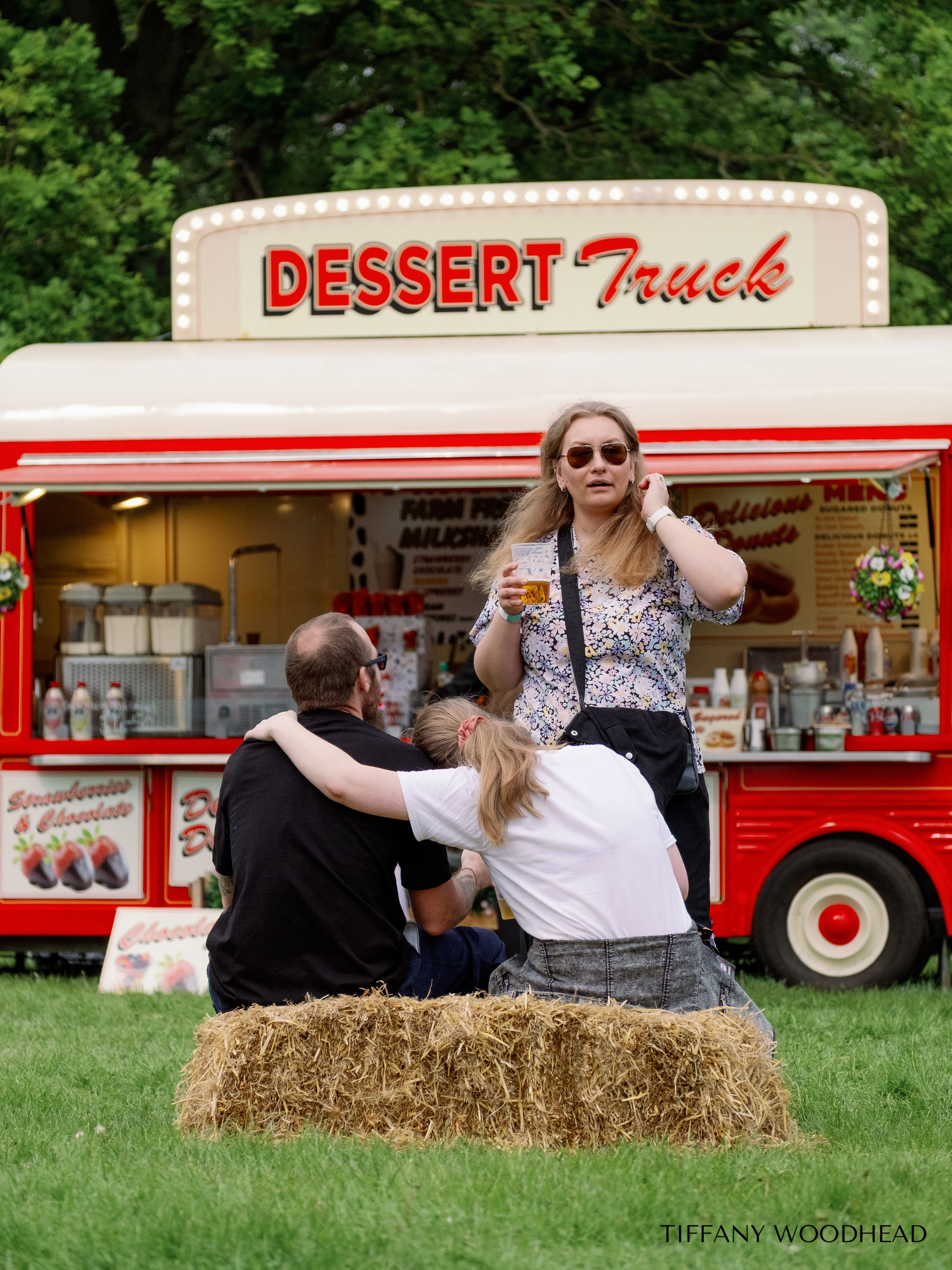 People sitting on a hay bale in front of a red dessert truck. One woman with long hair wearing sunglasses and floral shirt is standing, while two others are seated. The truck is decorated with bright lights and menu signs, surrounded by trees.