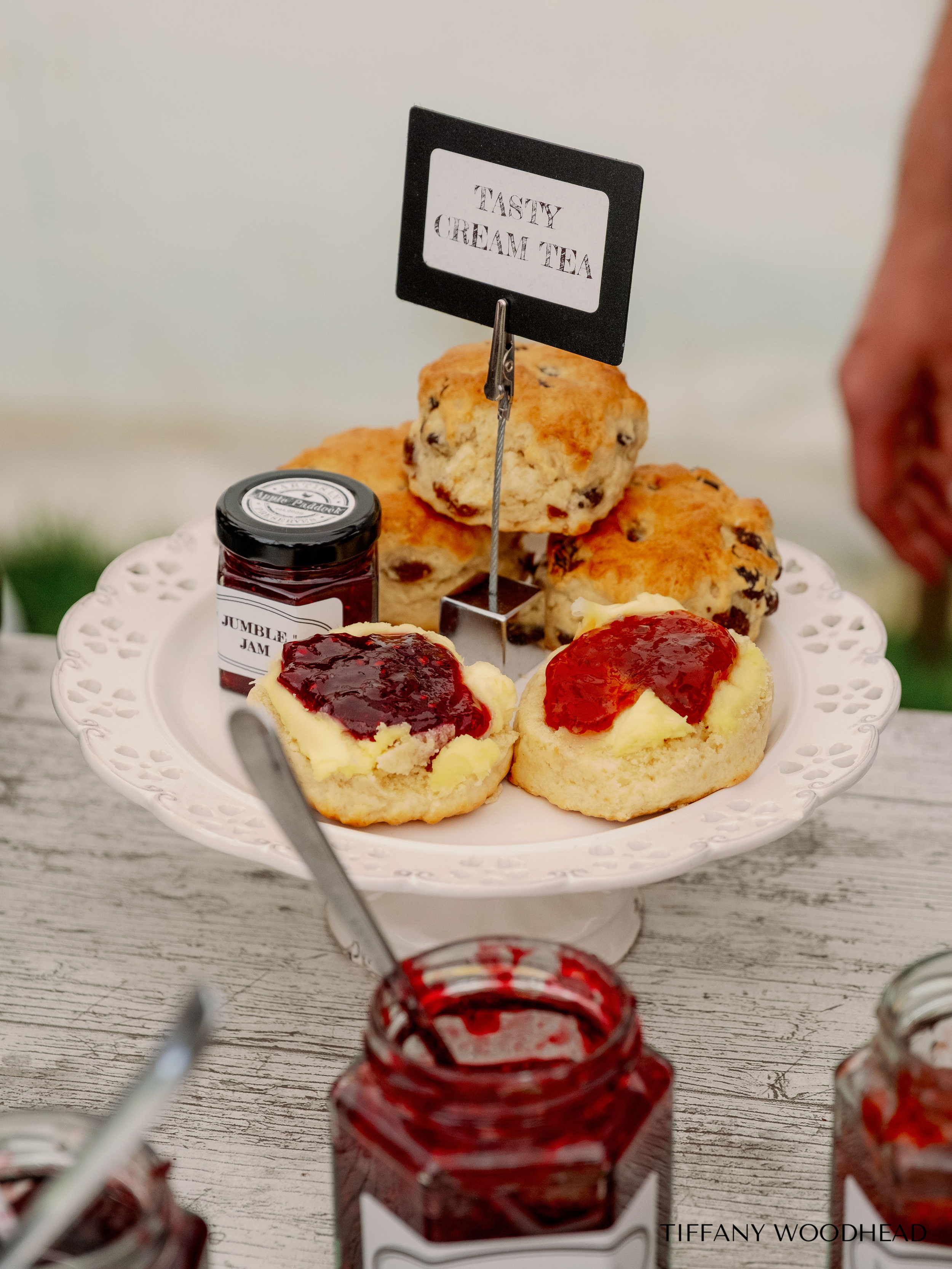 A plate holds two scones with jam and cream, along with a jar of jam; behind the plate are two more scones and a jar of jam, with a sign that reads 'Tasty Cream Tea' on a stand.
