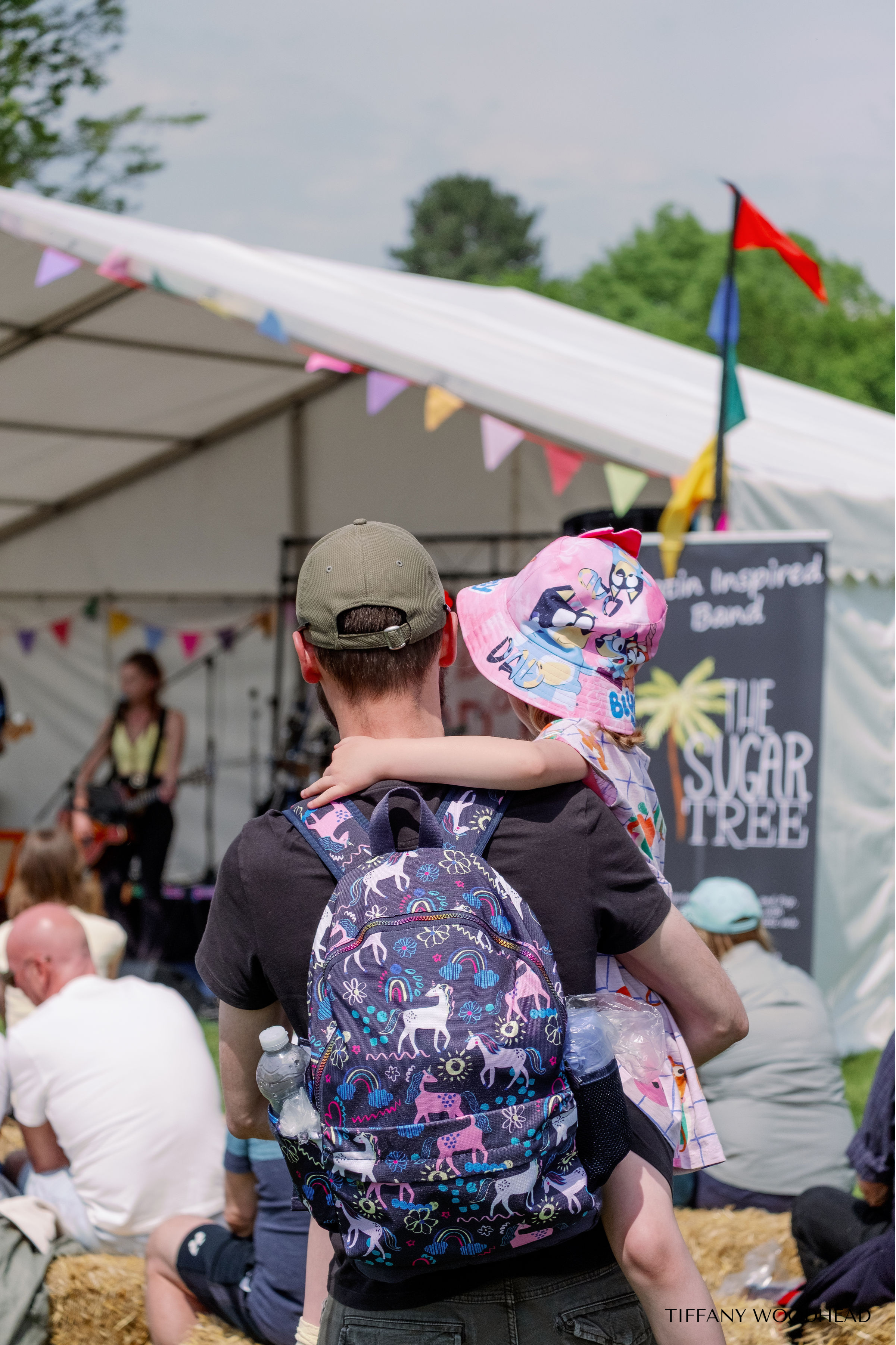 A man wearing a beige baseball cap and carrying a child on his shoulders, both facing away, at an outdoor event with a stage and banners in the background. The child is wearing a pink hat with cartoon characters and the man has a backpack with unicor