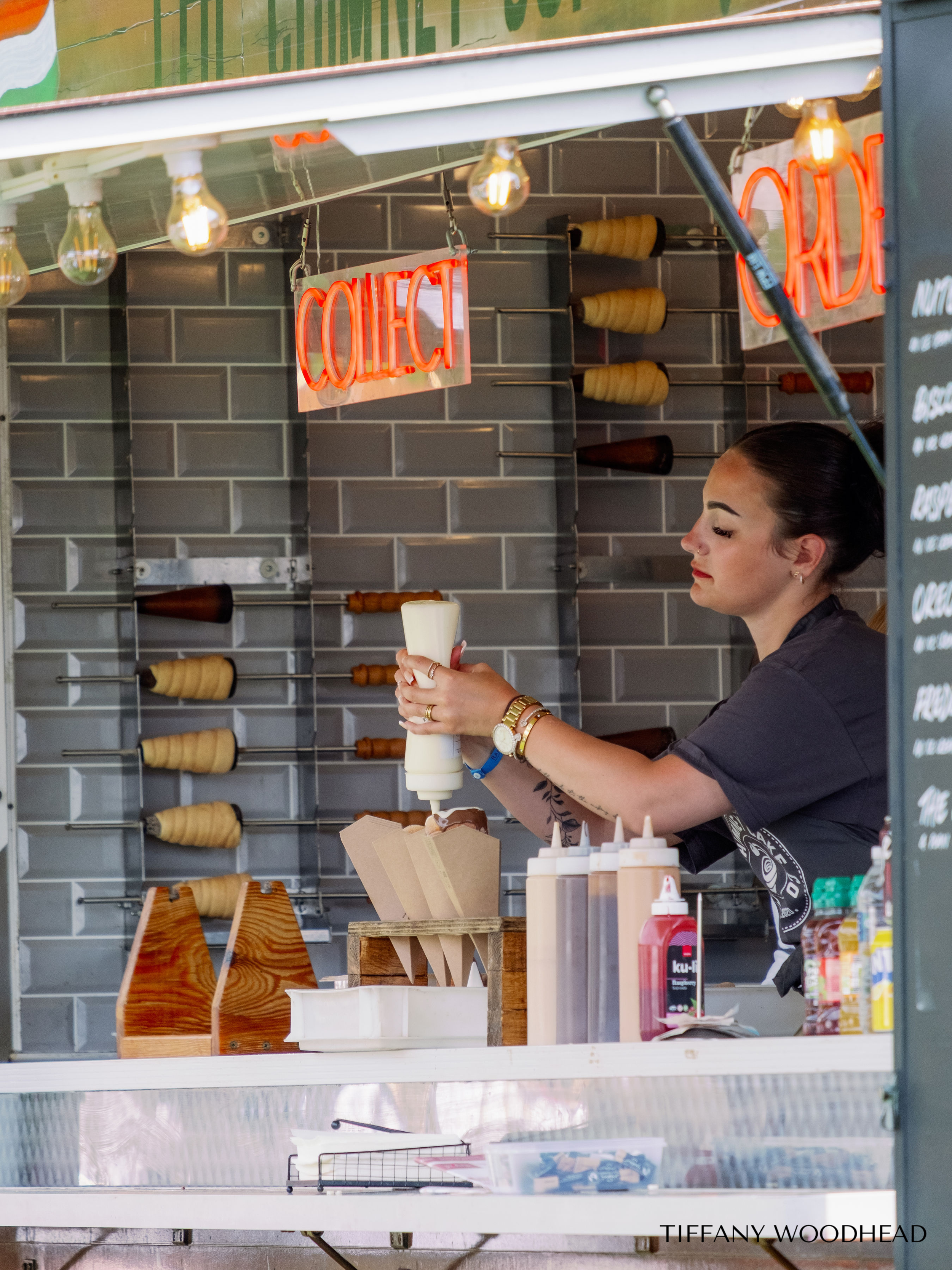 A woman making a soft-serve ice cream at a food stand, with various bottles of topping sauces on the counter, and a neon "CLOSED" sign hanging above.