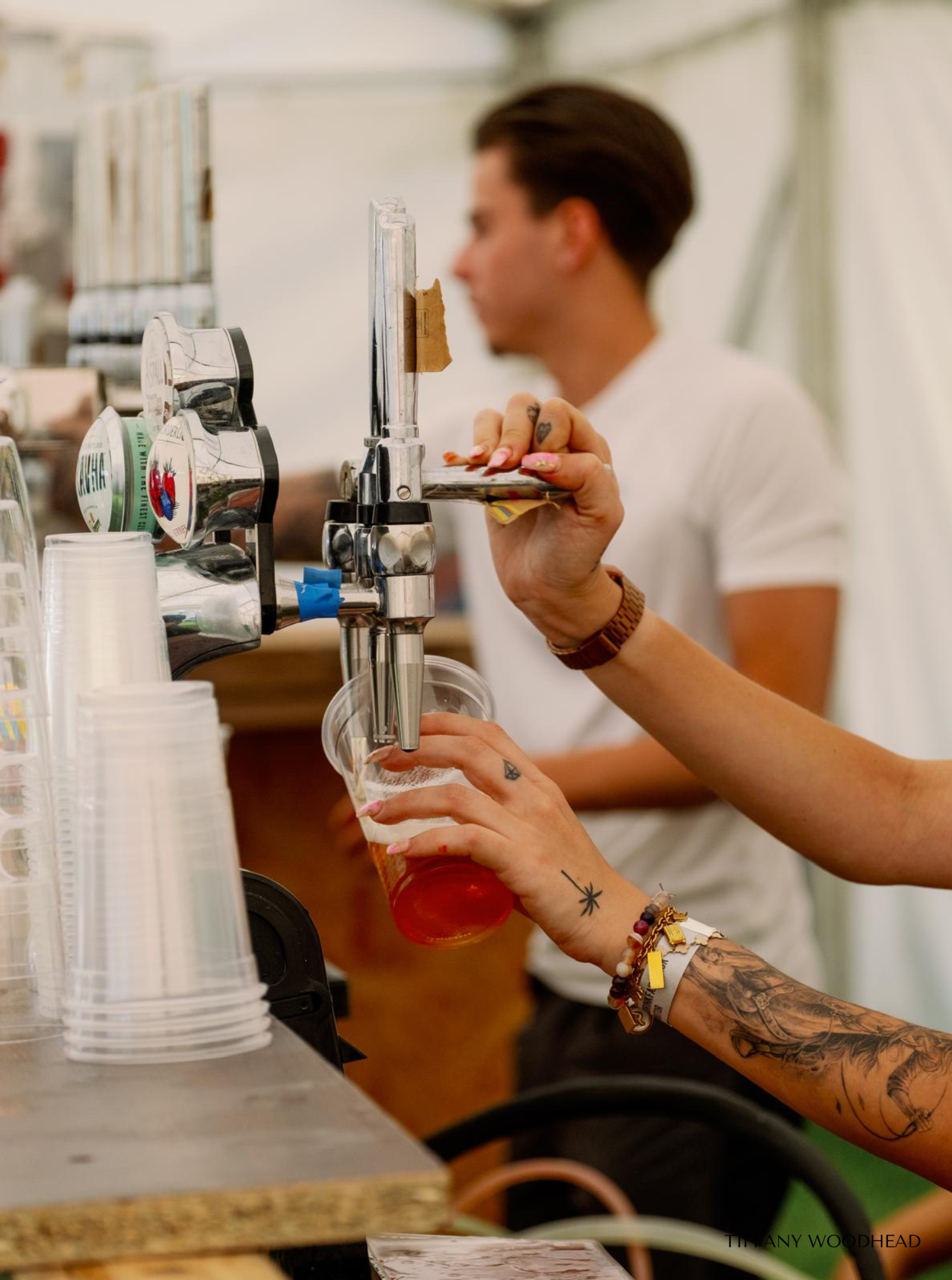 A person with tattoos on their arm is filling a plastic cup with beer from a tap at a bar or brewery. The person has jewelry and a wristwatch, and they are using both hands to hold the cup steadily.