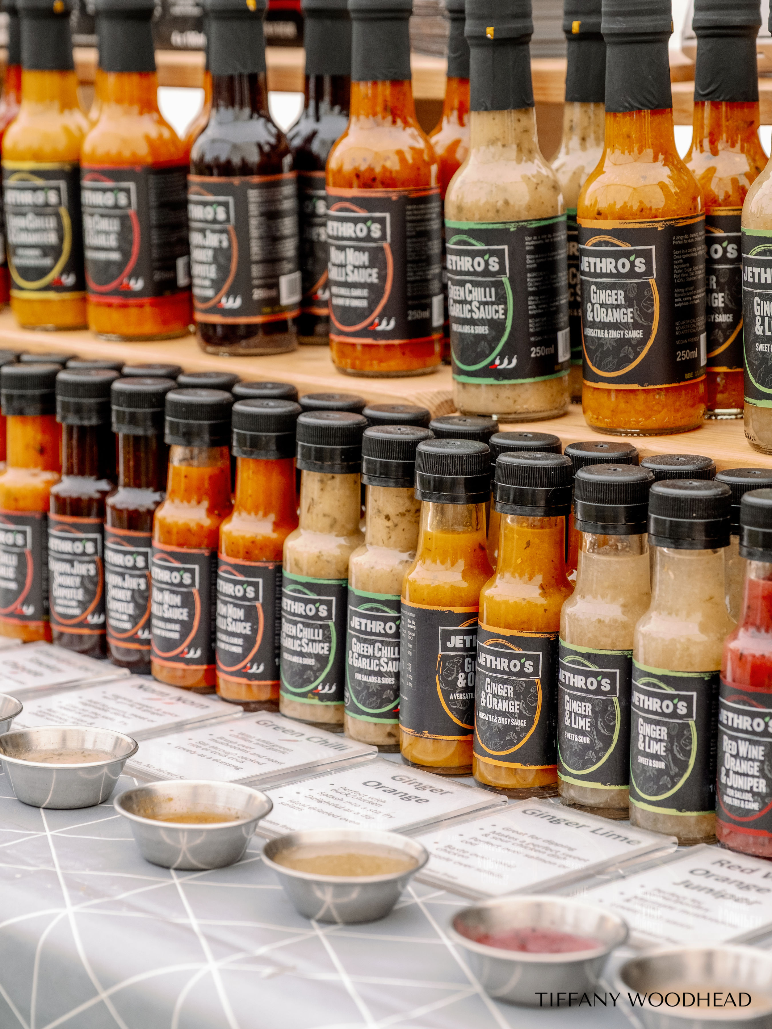Display of various hot sauces in glass bottles with black labels on a wooden shelf, with small metal bowls of sauces in front and menus below.