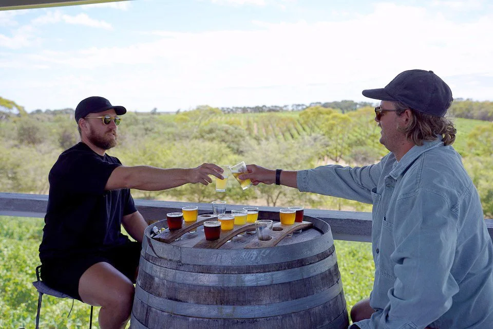 Two men toasting with Loophole Brewing Co. beer paddles at Cape Jaffa Wines' deck overlooking the vines — combining craft beer tasting with a laid-back vineyard setting.