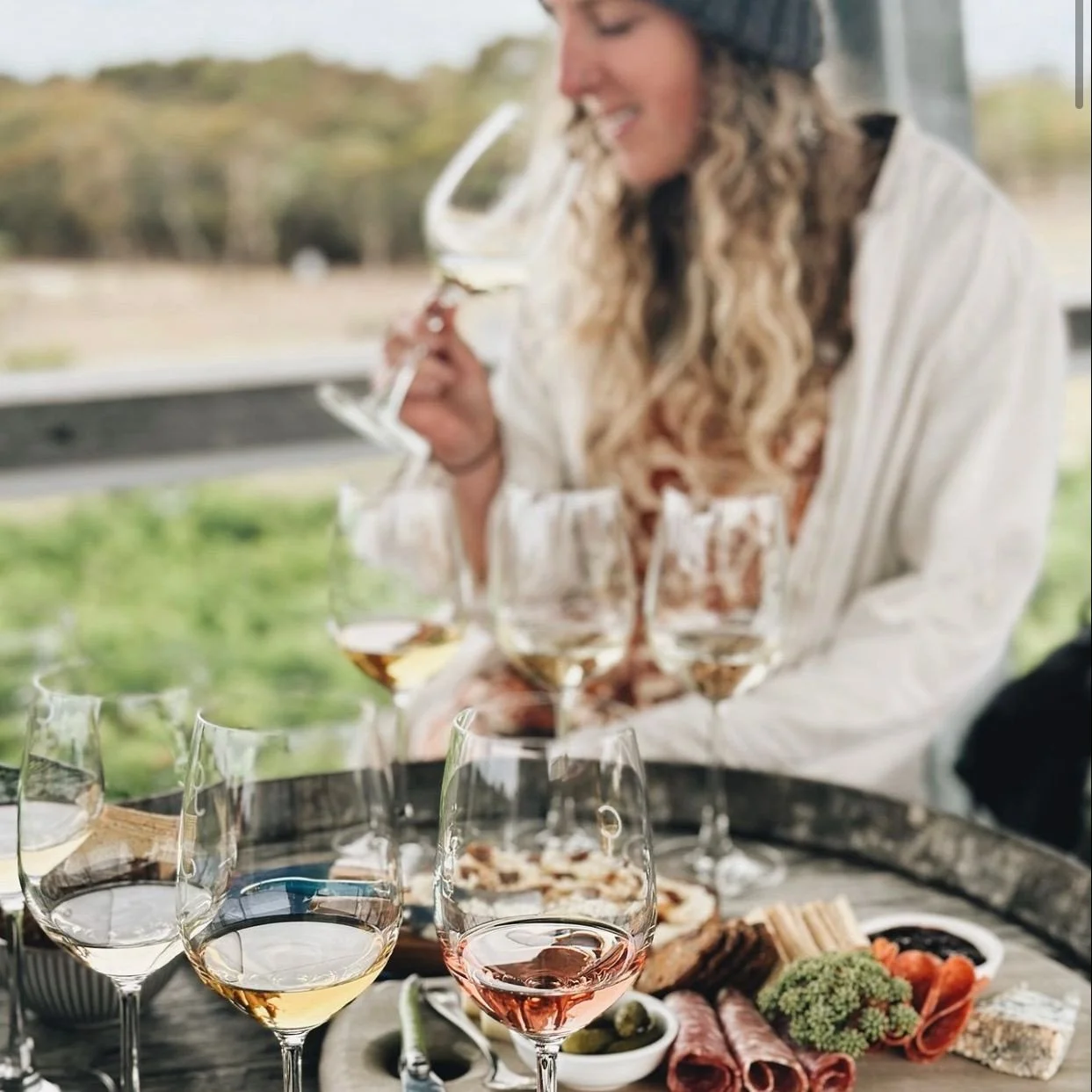 A woman holding a glass of Sauvignon Blanc sitting at a barrel underneath the covered deck over-looking over the vineyard — the image captures the beauty and stillness of Cape Jaffa Wines' rural wine country views.