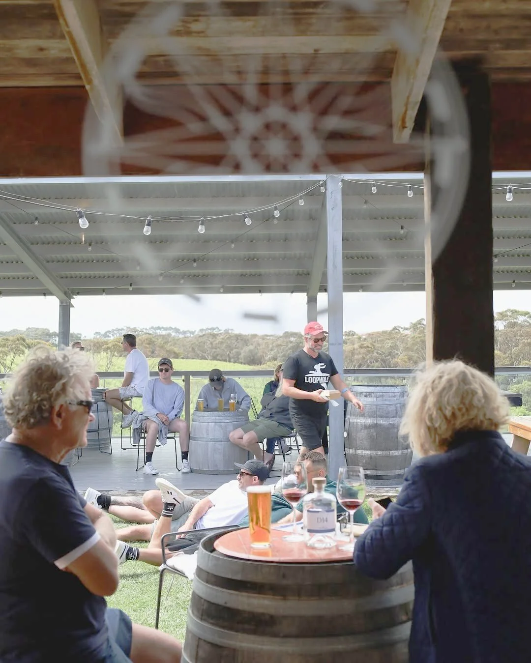 Window-framed view of guests lounging on the deck outside Cape Jaffa Wines cellar door — relaxed social scene overlooking vineyard rows on the coast.