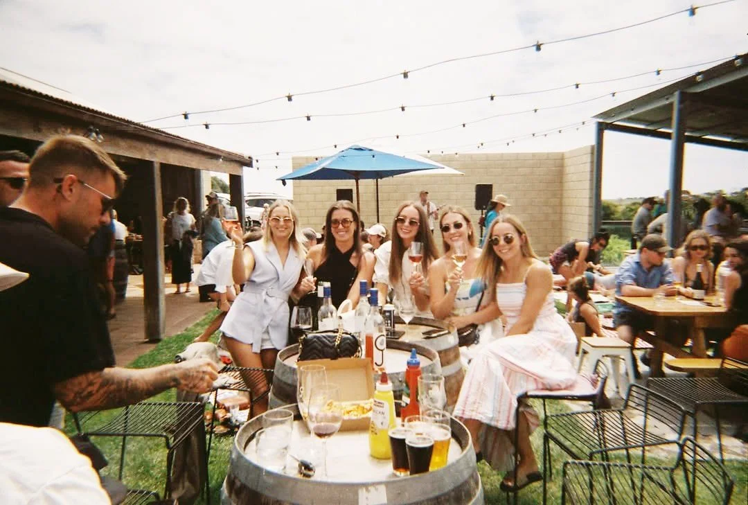 Group of friends gathered around wine barrels with drinks in hand at Cape Jaffa Wines' lawn bar — celebrating craft beer, summer vibes, and winery weddings near Robe.