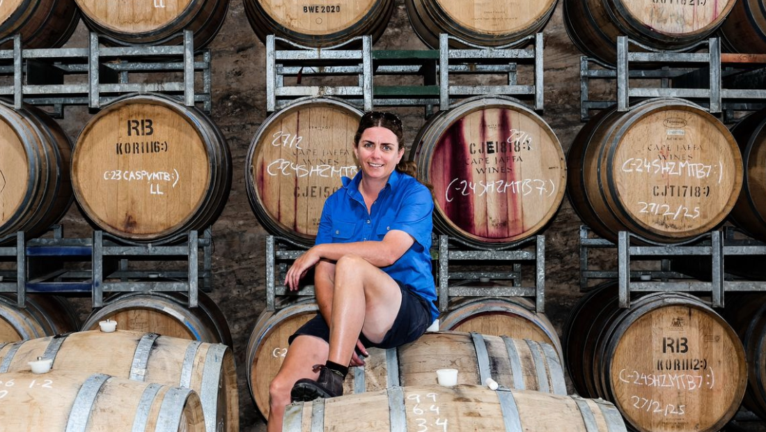 Anna Higgins Winemaker from Cape Jaffa Wines on the Limestone Cost in South Australia, nearby Robe; sitting on wine barrels in the winery. There is a backdrop wall of more wine barrels