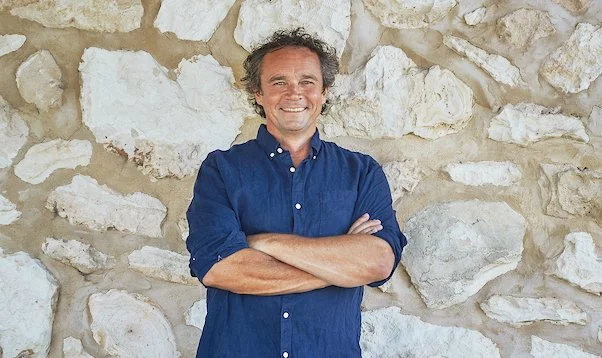 Derek Hooper, founder of Cape Jaffa Wines, standing in a blue shirt with arms crossed against a limestone wall; a leader in biodynamic winemaking near Robe.