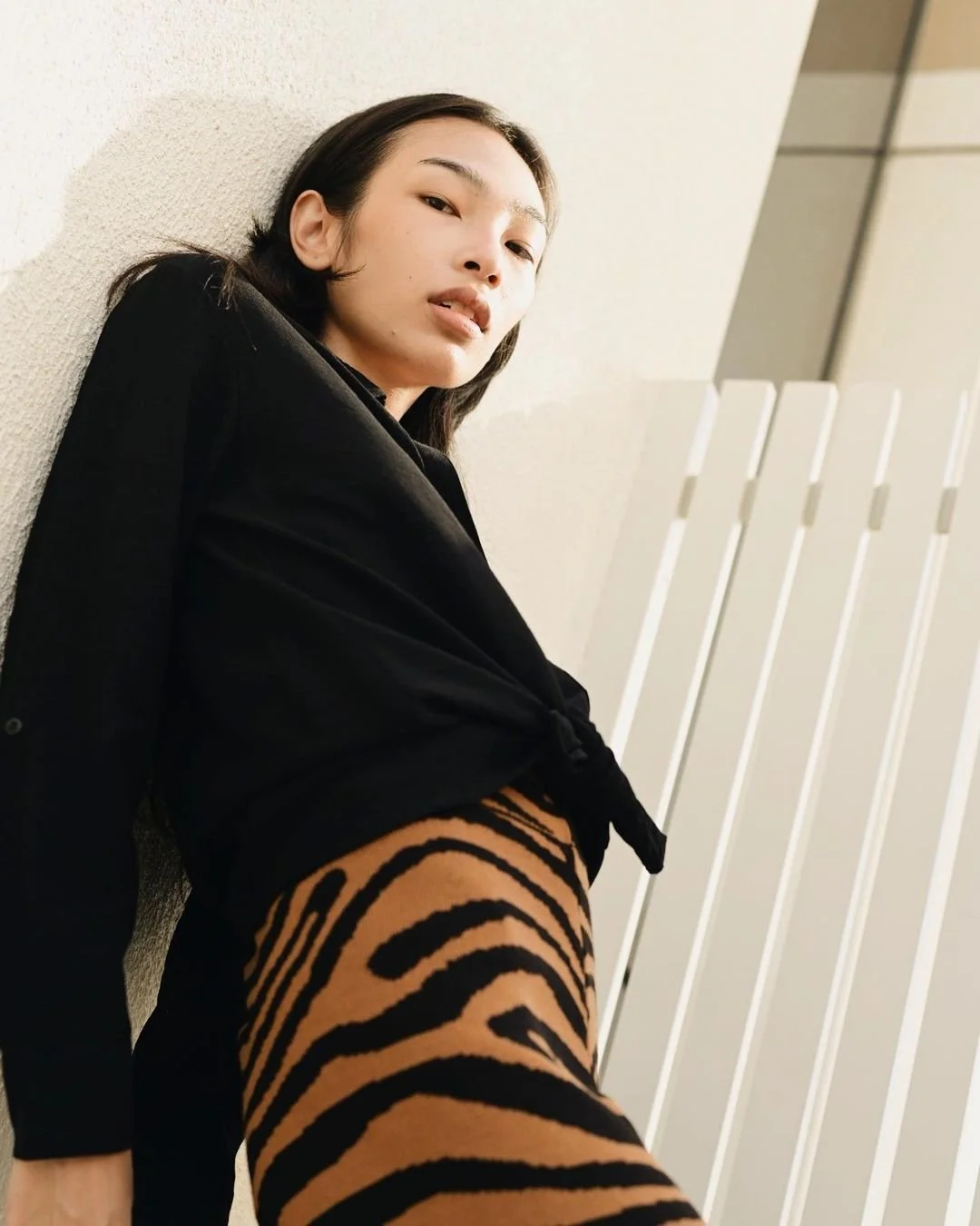 A woman leaning against a wall, wearing a black jacket and tiger-striped tights, looking into the camera.