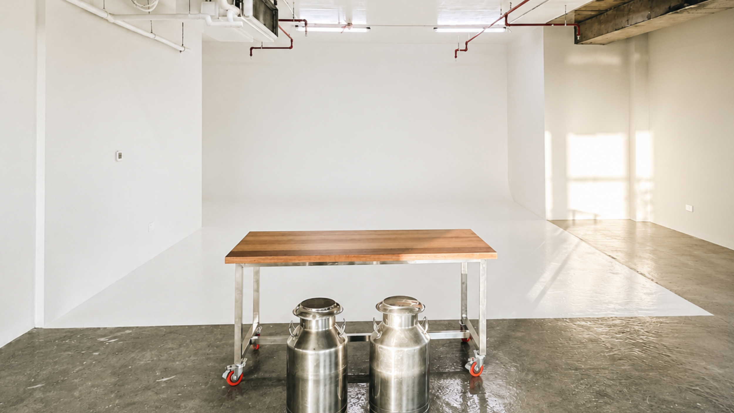 Empty room with white walls, a wooden table on a wheeled cart, and two metal milk cans on the floor. There is a window with sunlight streaming in, exposing the unfinished ceiling with visible pipes and beams.