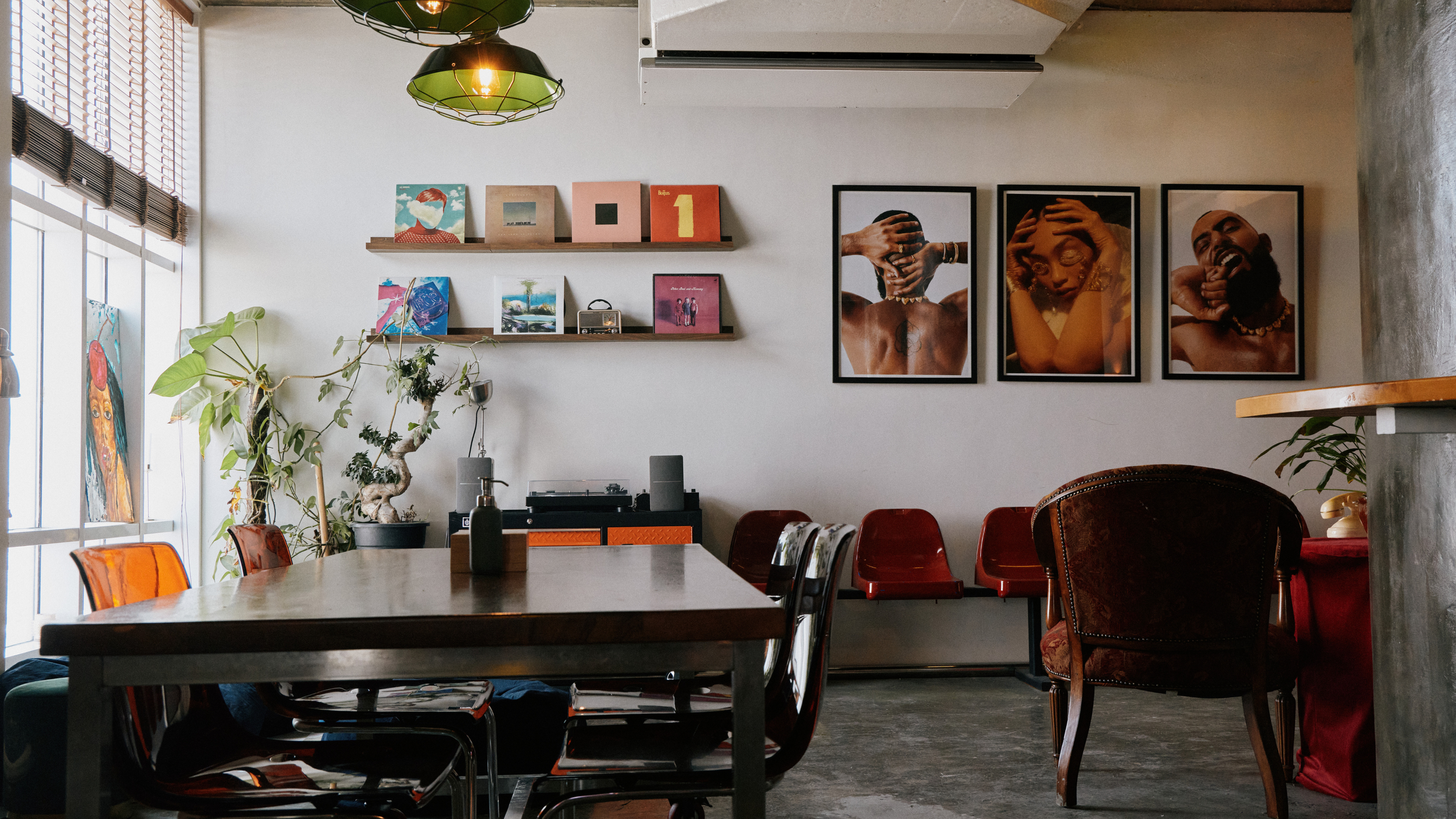 Interior of a room with a long dining table, surrounded by colorful chairs; wall decorations including vinyl records, paintings, and framed photos of people with tattoos and jewelry; potted plant in corner near window, and a vintage radio on a shelf.