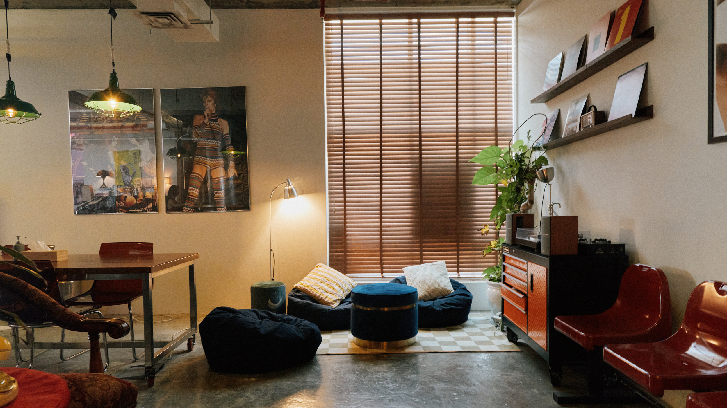 Cozy living room with wooden blinds on large window, framed photographs, a small plant, colorful seating cushions, vintage record player, and modern pendant lights.