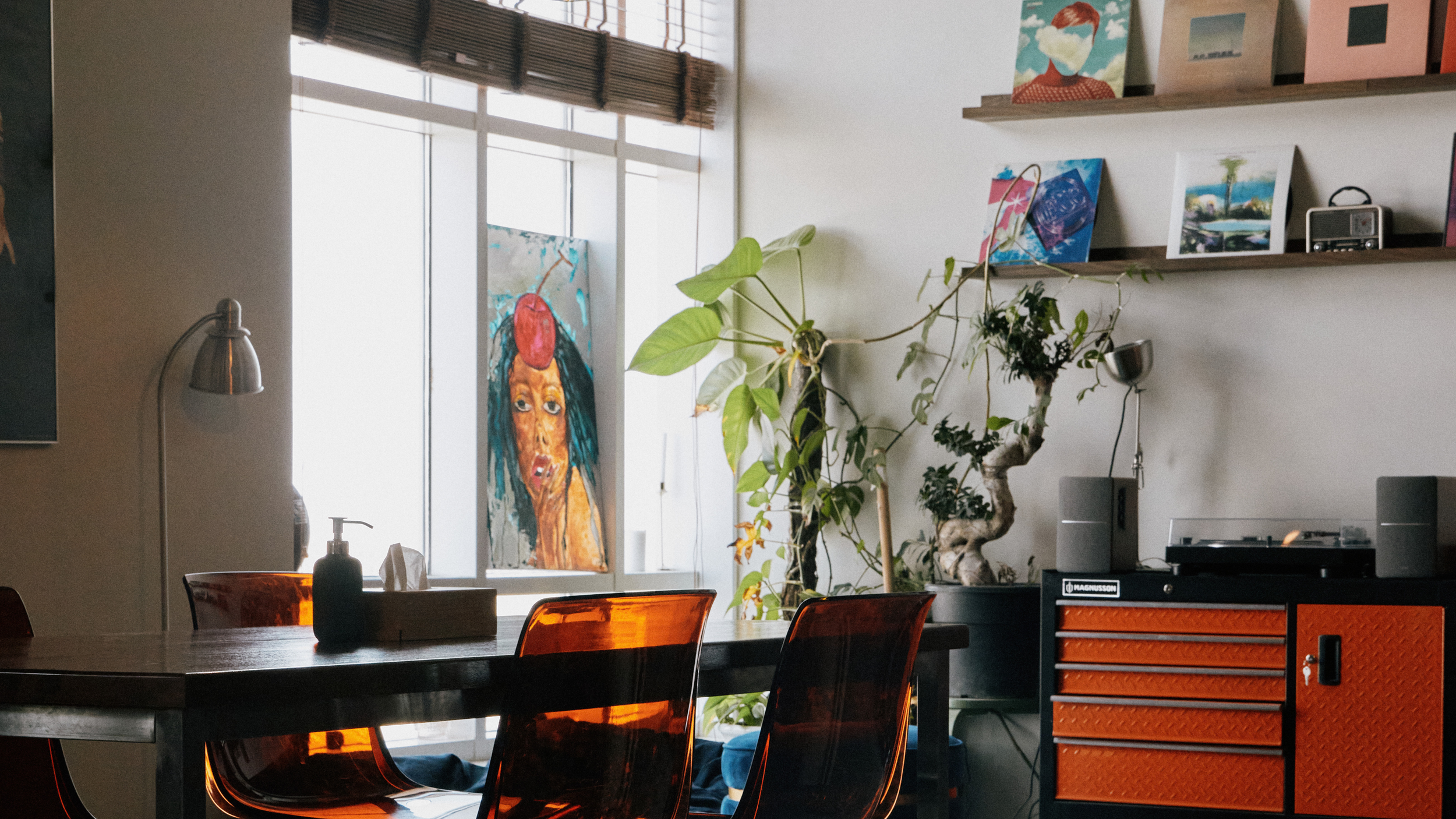 Interior of a modern room with a large window, a colorful painting of a woman with blue hair and a red object on her head, green houseplants, a wooden shelf with art and decorative items, and orange chairs around a gray table.