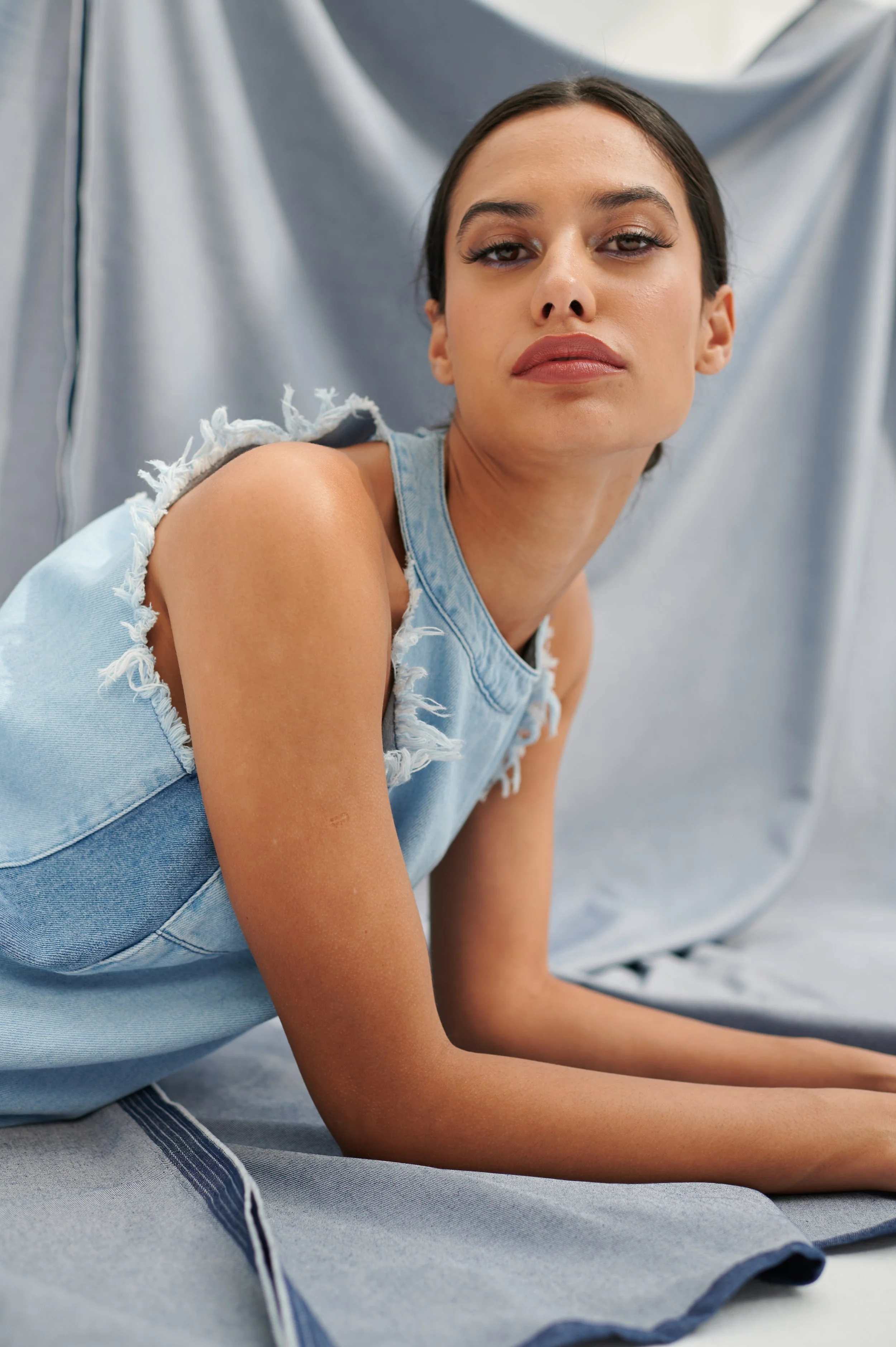 A young woman with dark hair and makeup sitting against a silver fabric backdrop, wearing a sleeveless frayed denim top.