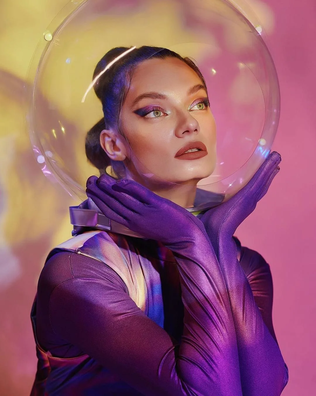 A woman in space-themed costume with purple gloves and a clear helmet, posing with her hands on her neck against a colorful, abstract background.